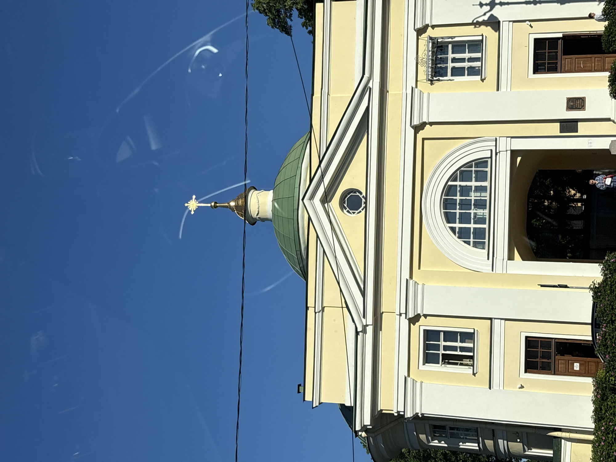 The image displays the Church of the Holy Spirit, situated within the Alexander Nevsky Lavra in Saint Petersburg, Russia. The building features a pale yellow stucco facade accented with white neoclassical architectural elements, including prominent cornices, pilasters, and window frames. A large, dark green dome with a light-colored drum rises above the central section, surmounted by a smaller, gilded onion dome topped with a golden, eight-pointed Orthodox cross. Below the dome, a triangular pediment contains a circular oculus window.
At the ground level, a broad, arched entryway provides access to a dark interior passage. To the immediate left of this arch, a dark brown wooden door is partially ajar, revealing a darkened interior. Another identical, partially ajar dark brown wooden door is positioned to the right of the central arch. Above the right-hand door, a rectangular window with white muntins is visible, fitted with an ornate black metal canopy. Above the left-hand door, another rectangular window is present, with its upper sash open. Small, dark plaques with unreadable text are affixed to the wall surfaces to the right of the central arched entry.
The sky above is clear and intensely blue, with multiple faint, elongated streaks and reflections consistent with an image taken through a glass pane, such as a car window. A single black utility cable spans horizontally across the upper mid-ground of the scene. Lush green foliage, primarily dense bushes, borders the base of the building, partially obscuring its lower sections. Additional green tree foliage is visible on the far right side of the frame, behind the structure. Sunlight illuminates the building from the left, casting a distinct shadow, likely from a nearby lamppost, onto the building's right side.
Two individuals are present: one person, dressed in light-colored clothing and carrying a light-colored bag, stands partially visible within the left segment of the main arched entryway. A second individual, wearing a light-colored shirt and dark trousers, is partially visible on the far right edge of the frame, largely obscured by the foreground bushes.
