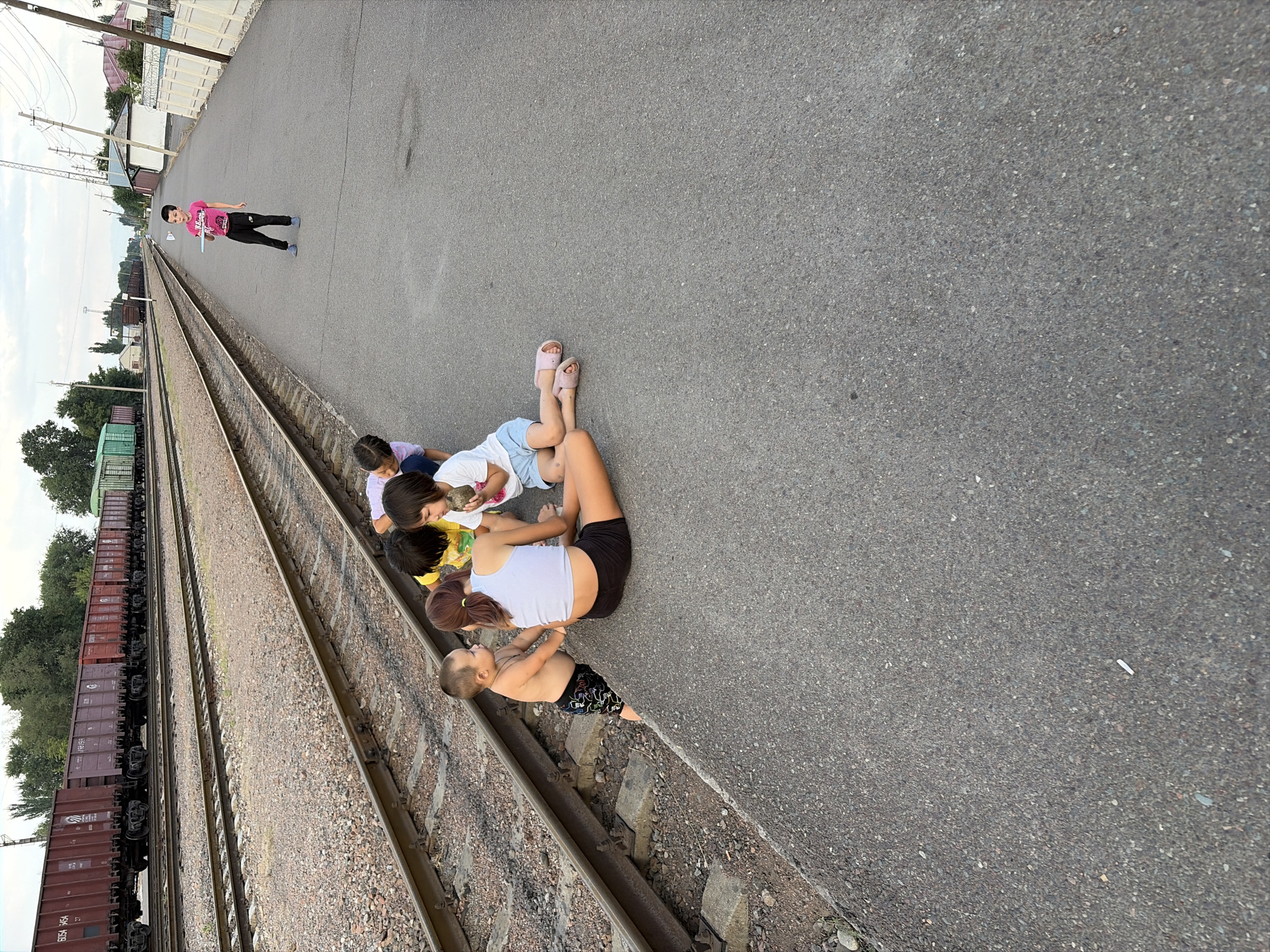 A group of children are gathered on a paved surface next to railroad tracks in what appears to be a station or industrial area. The scene is captured from a high angle, with the camera rotated.
There are at least five children visible. One child, wearing a pink t-shirt and black pants, stands slightly apart from the others, holding a white object. The remaining children are sitting or lying on the ground, interacting with each other. One child, in a white tank top and black shorts, is on her back, with her legs bent. Another child, wearing a yellow shirt, is leaning over a smaller child who is lying down. Two other children, appearing to be girls, are also in the group, looking down.
The setting is an outdoor area with two sets of railroad tracks running parallel to each other. A long train with numerous brown freight cars is visible on the left track. The ground is a mix of asphalt and gravel. In the background, there are some buildings, fences, and trees, suggesting an urban or semi-urban environment.
The lighting suggests it is daytime, possibly late afternoon or early evening, as the light is somewhat diffused. The weather appears to be fair. There are no overt expressions of strong emotion, but the children seem engaged with each other, possibly playing or resting.
No visible text is present on the clothing of the children. Some of the train cars have markings, but they are not legible in the image. The location context provided is Kant, Kyrgyzstan.