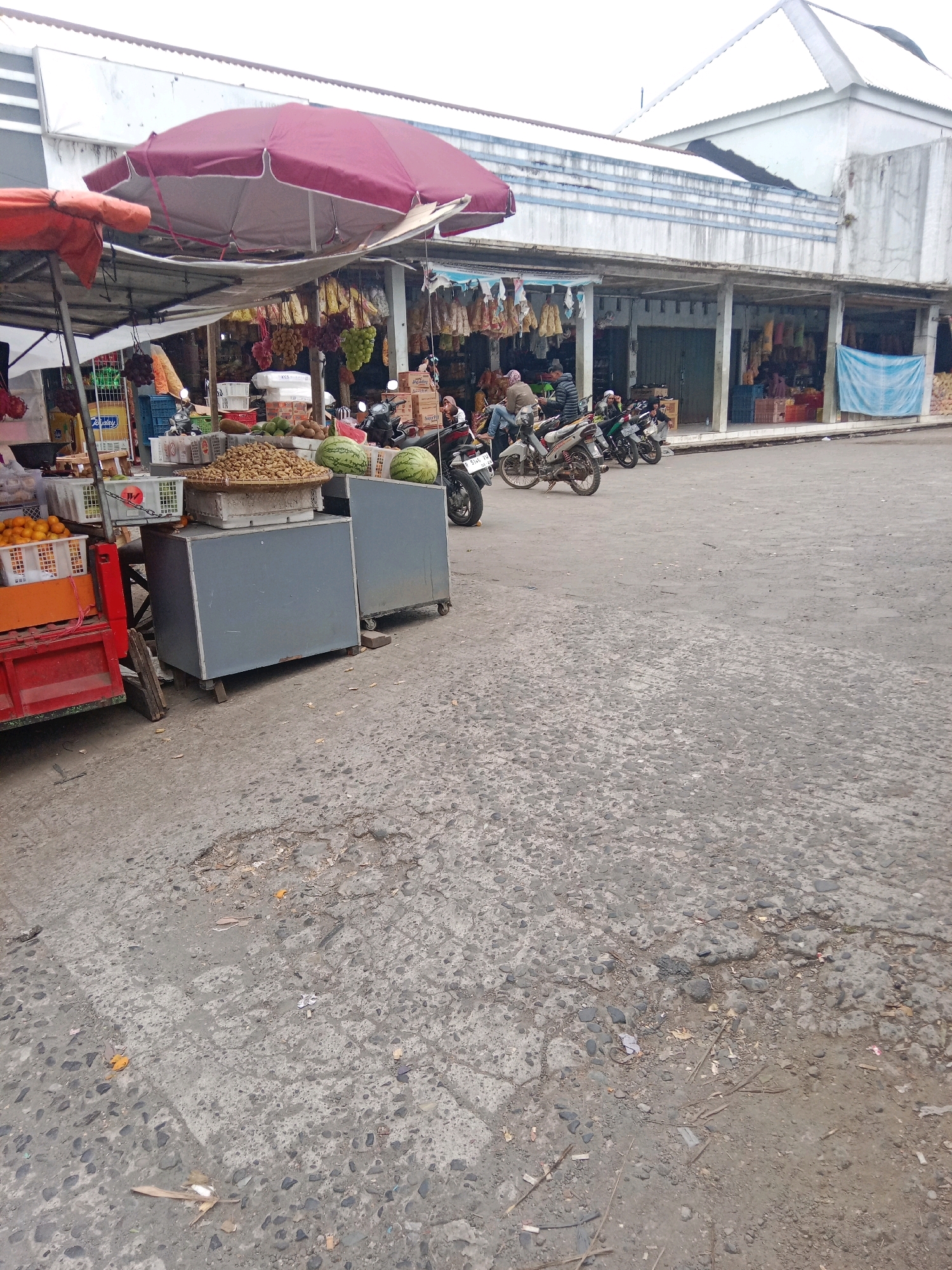This is a vertical, eye-level, medium shot of an outdoor market. The market appears to be in the city of Batur, Indonesia. In the foreground and to the left, a red cart is visible, laden with an orange crate filled with oranges. Next to the cart are two gray, wheeled cabinets. On top of the left cabinet is a woven basket filled with small, brown objects, possibly nuts or seeds. Two watermelons sit on top of the right cabinet. Above the fruit displays, a large, burgundy umbrella is propped up, casting a shadow. In the background, several motorcycles are parked. People are visible in the background, some seated on the motorcycles and others standing near market stalls. The stalls themselves are lined up under a covered walkway with concrete pillars. Various goods, including dried foods and textiles, hang from the stalls. The ground is paved with rough concrete, showing signs of wear and tear, with scattered debris. The overall scene suggests a bustling, yet somewhat weathered, local market.