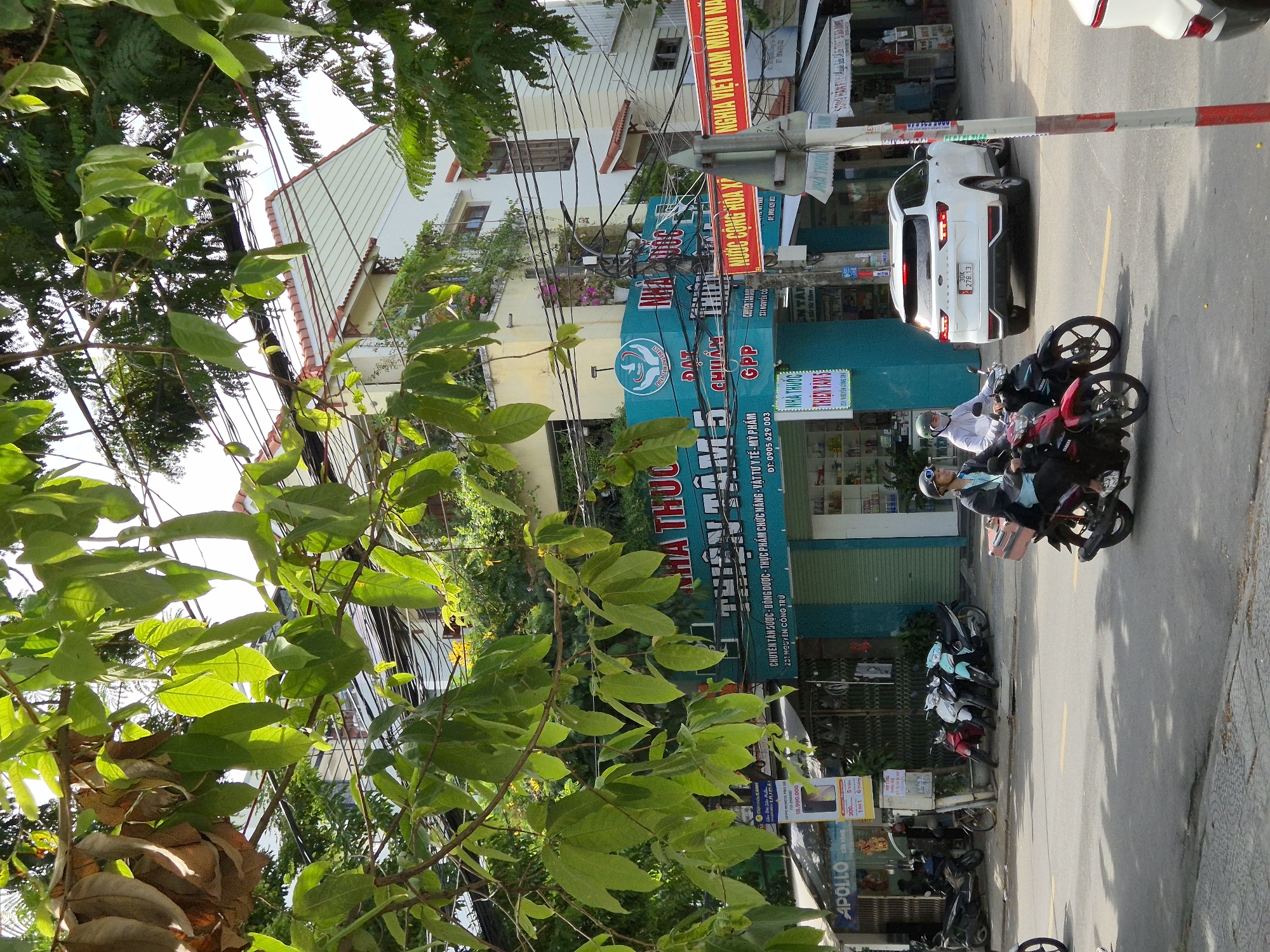 The image depicts a street scene in Da Nang, Vietnam, on a sunny day. The foreground shows a street with a few motorcycles and a white SUV. Two people are riding on a motorcycle in the foreground, one driving and the other as a passenger. Behind them, parked motorcycles are visible. Across the street, buildings with businesses are lined up. One building has a turquoise facade with text in Vietnamese, appearing to be a pharmacy or medical supply store. Another building is white with a red-tiled roof. Overhead, a tangle of electrical wires and lush green foliage from trees frame the upper portion of the image. A red banner with Vietnamese text is visible on a pole in the middle ground. The overall impression is of a bustling urban environment.