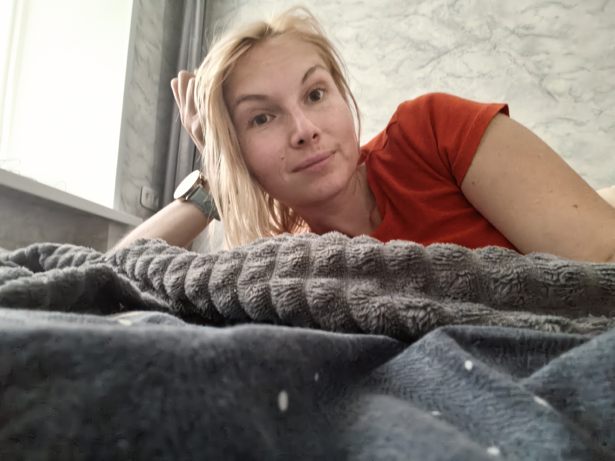 A blonde woman with light skin is lying in bed. She is wearing an orange t-shirt. Her right arm is bent and her hand is resting on her head. She has a watch on her left wrist. The background is a marble-patterned wall and a window with grey curtains. The bed is covered with dark grey blankets. The scene appears to be indoors, possibly in a bedroom. There is no specific information to indicate the location in the city of Sterlitamak, Russia.