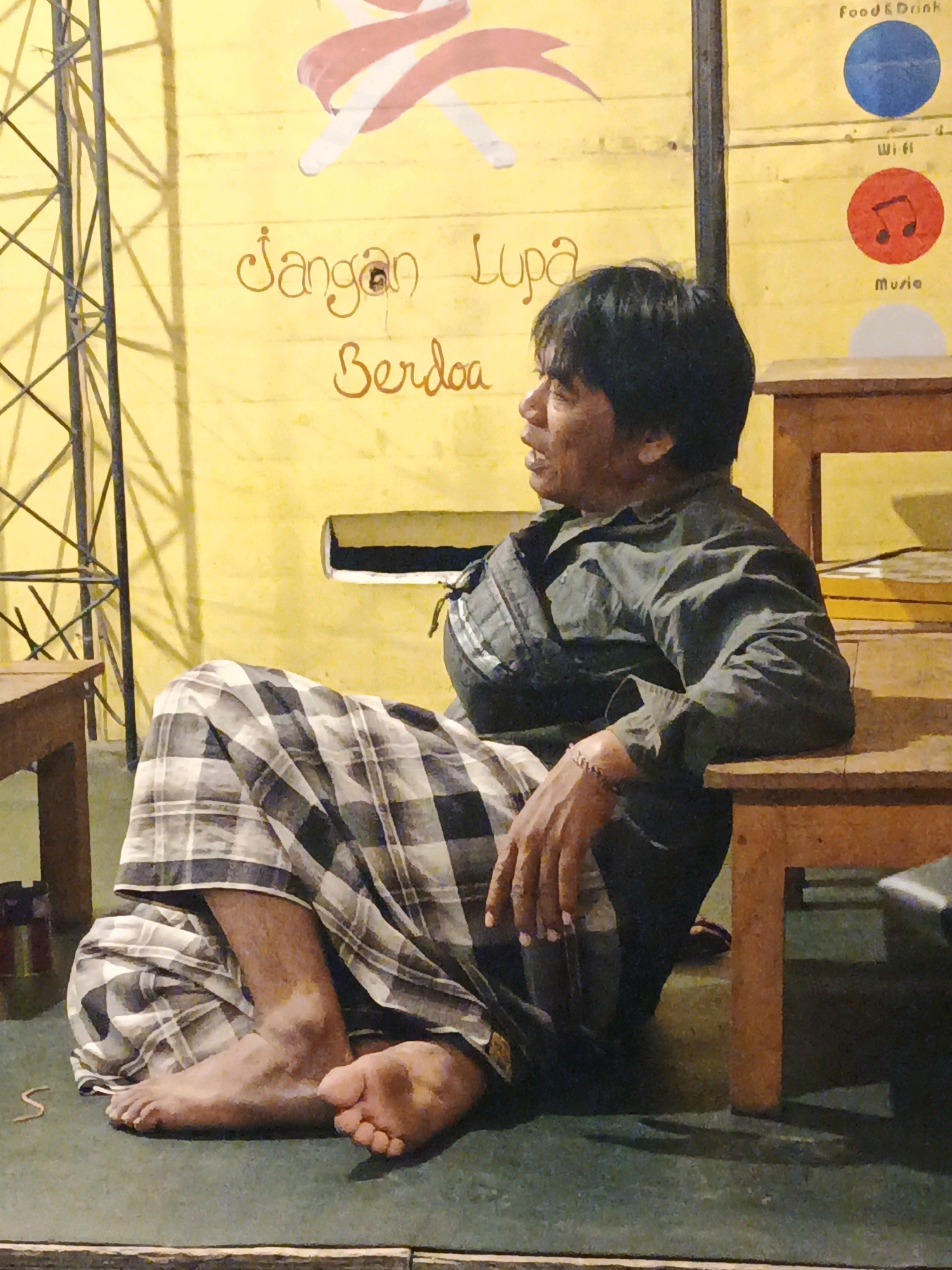 A man in a plaid sarong and dark green long-sleeved shirt is sitting on a green floor next to a wooden bench in what appears to be an outdoor or semi-outdoor establishment. He is looking to his right, with his mouth slightly open, as if speaking. The man's left arm is resting on his lap, and his right arm is bent, with his hand resting on his right knee. His feet are visible, with his left foot flat on the floor and his right foot slightly raised and angled. The background is a yellow wall with writing in Indonesian that says "Jangan Lupa Berdoa," which translates to "Don't Forget to Pray." There are also circular icons on the wall with text labels: "Food & Drink," "Wifi," and "Music." To the left of the man, a portion of a metal tower structure is visible. Wooden tables are present in the scene. The lighting suggests it is likely daytime, with shadows cast by the man and the furniture. The location is identified as Ketegan, Indonesia.