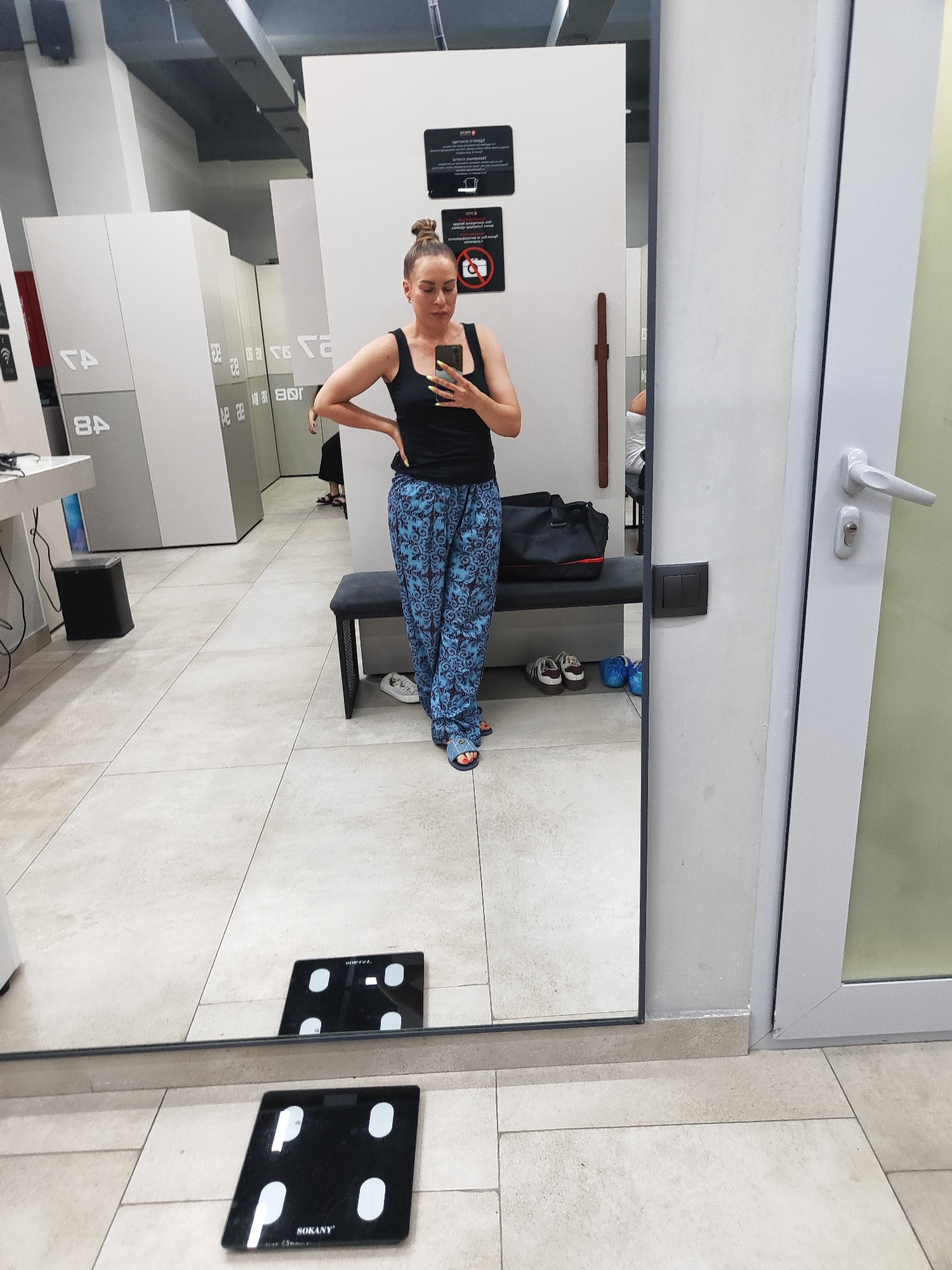 A woman stands in a locker room, taking a mirror selfie. She is wearing a black tank top and blue patterned pants, with her hair pulled up in a bun. She is holding a smartphone in her right hand and has her left hand on her hip. In the background, there are lockers, a bench, and a scale on the floor. The scene appears to be well-lit, suggesting it is daytime. The woman's expression is neutral. The location is identified as Almaty, Kazakhstan. Visible text on the scale reads "SOKANY". Signs are present on the lockers, but the text is not clearly legible.