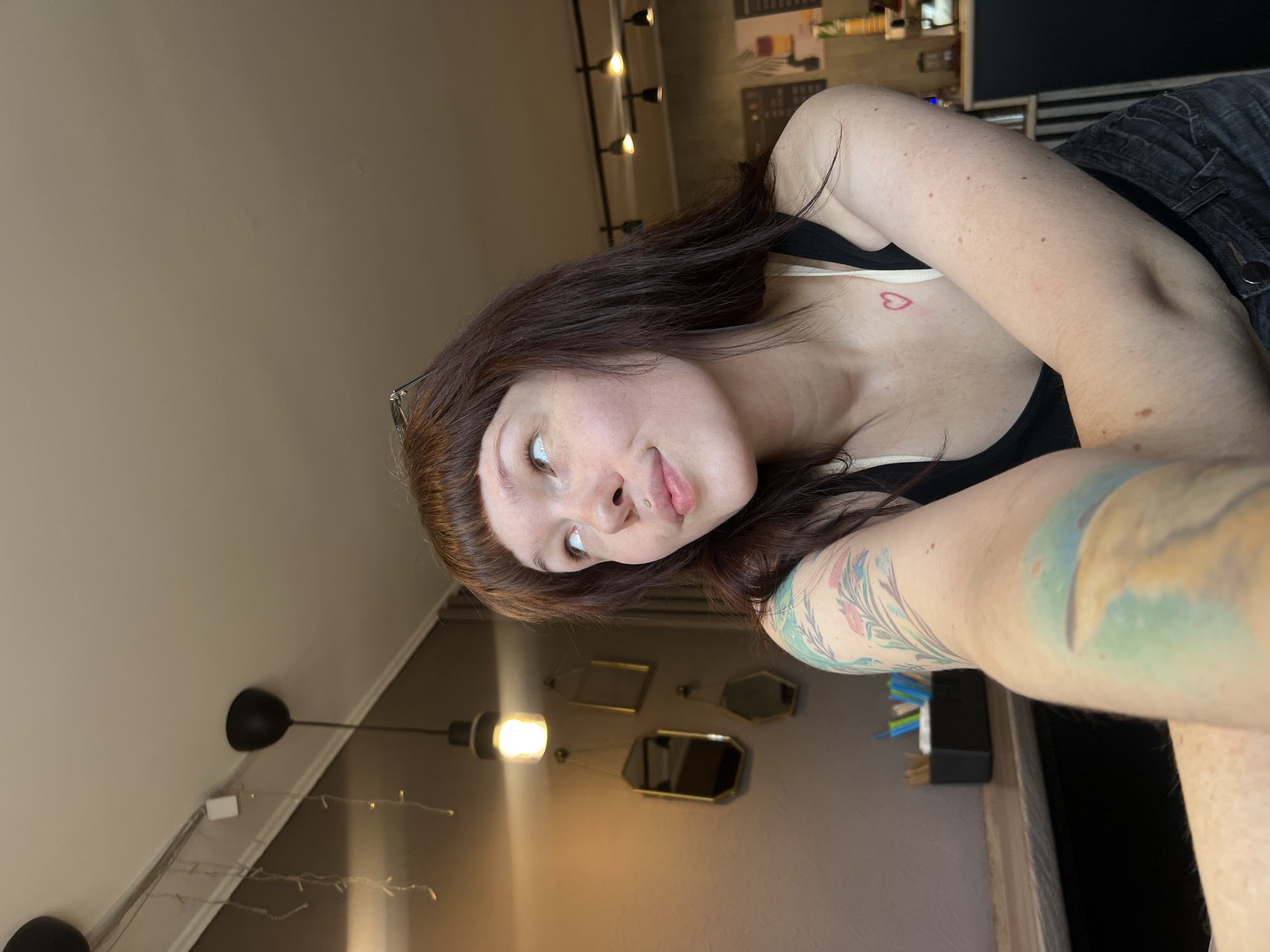 The image shows a young woman with brown hair, fair skin, and a light pink tint to her lips, looking towards the camera with a slightly playful expression. She has a small heart tattoo on her chest and a large, colorful tattoo on her arm. She is wearing a black tank top and denim shorts. The setting appears to be indoors, with a dimly lit room that has warm-toned walls. Several light fixtures are visible on the ceiling, and decorative mirrors are hung on the wall behind her. The lighting suggests it could be evening or a room with minimal natural light. There are no visible weather cues. The woman's pose and expression suggest a casual, perhaps posed, selfie.