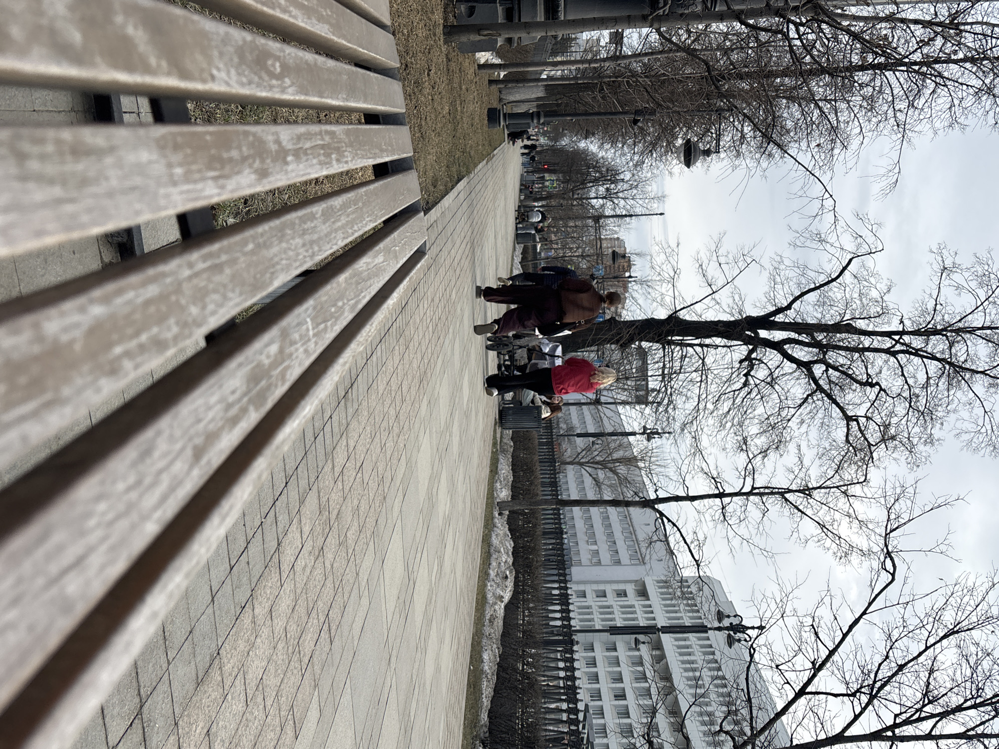 A long, grey, wooden park bench dominates the left foreground of the image. The perspective is low, looking along the length of the bench towards a paved pathway. The pathway is lined with trees, most bare of leaves, suggesting late autumn or early spring. Two people are walking along the path, one wearing a brown coat and the other a bright red jacket. A white building with multiple windows stands in the background. The sky is overcast. The image was taken in Perm, Russia.