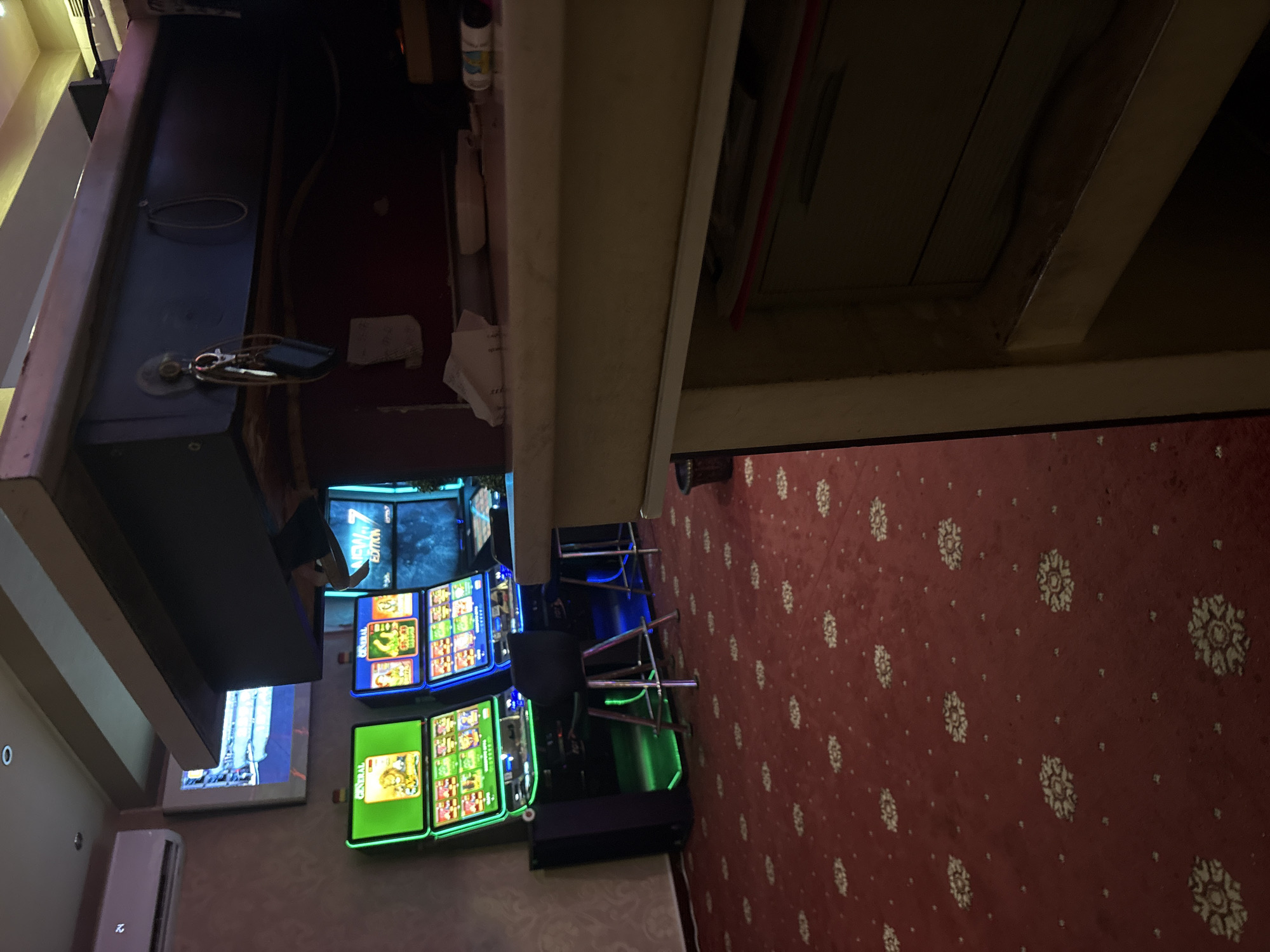 In Karlovo, Bulgaria, inside what appears to be a dimly lit casino or entertainment establishment, three slot machines are visible on the left side of the frame, with their screens illuminated. The leftmost machine displays a green interface with a lion graphic at the top, and the second machine from the left features a blue interface with a crocodile graphic. The third machine is partially obscured by the second and displays a blue screen with the text "NEW EDITION." Red carpeting with a repeating floral pattern covers the floor. Two black bar stools with silver legs are positioned in front of the slot machines. Above the slot machines, a television screen displays an image that is difficult to discern. A dark, overhanging structure, possibly a bar or counter, extends from the top of the frame, with keys and a loop hanging from it. The right side of the image is dominated by the foreground of a light-colored counter or service area.