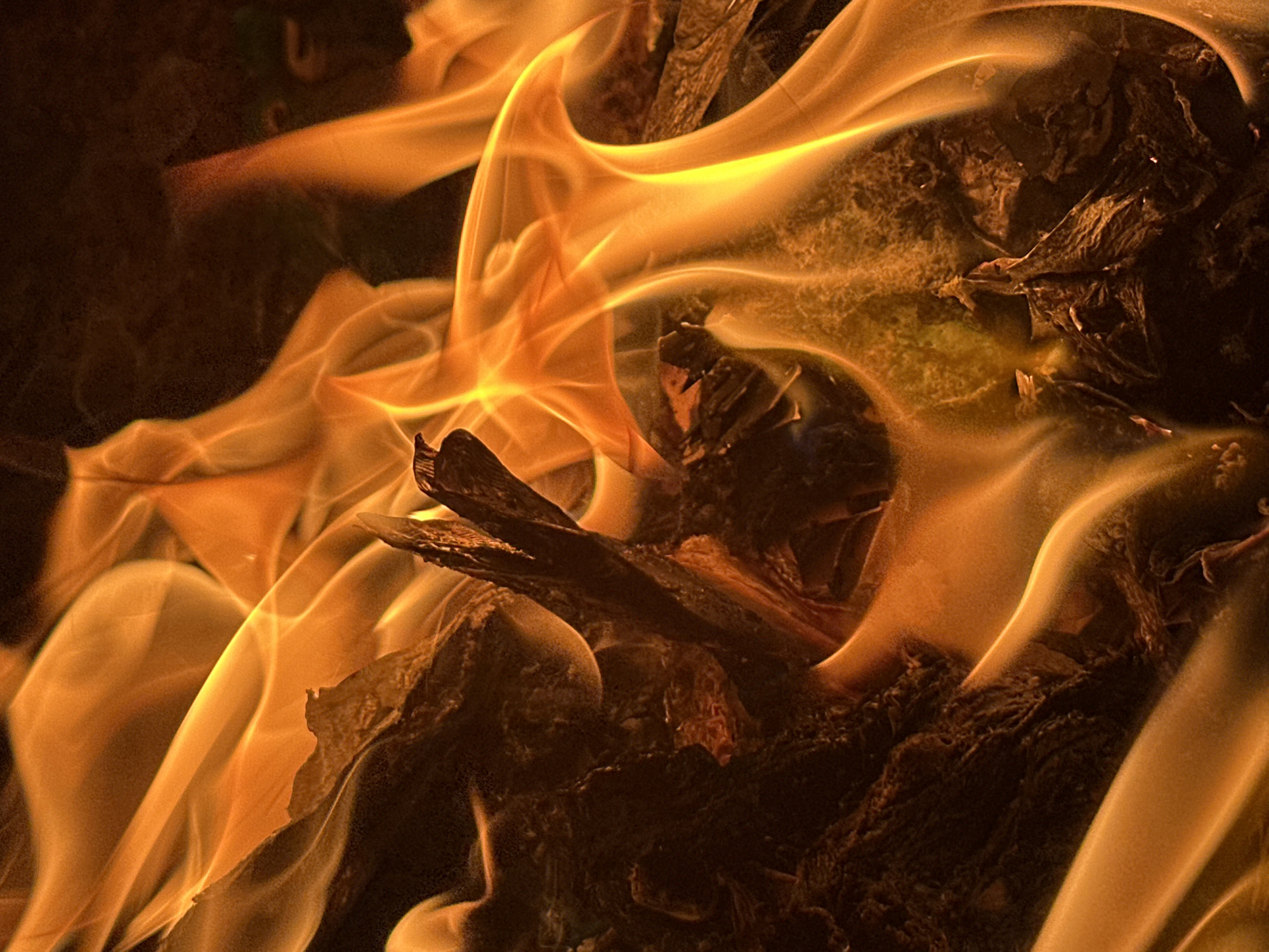 The image shows a close-up view of a fire. Flames, varying in color from yellow to orange, are prominent and appear to be engulfing pieces of wood. The wood is dark and charred. The environment is dark. No people are visible. No specific location details such as Bar Gada, Nigeria, can be verified from the image.
