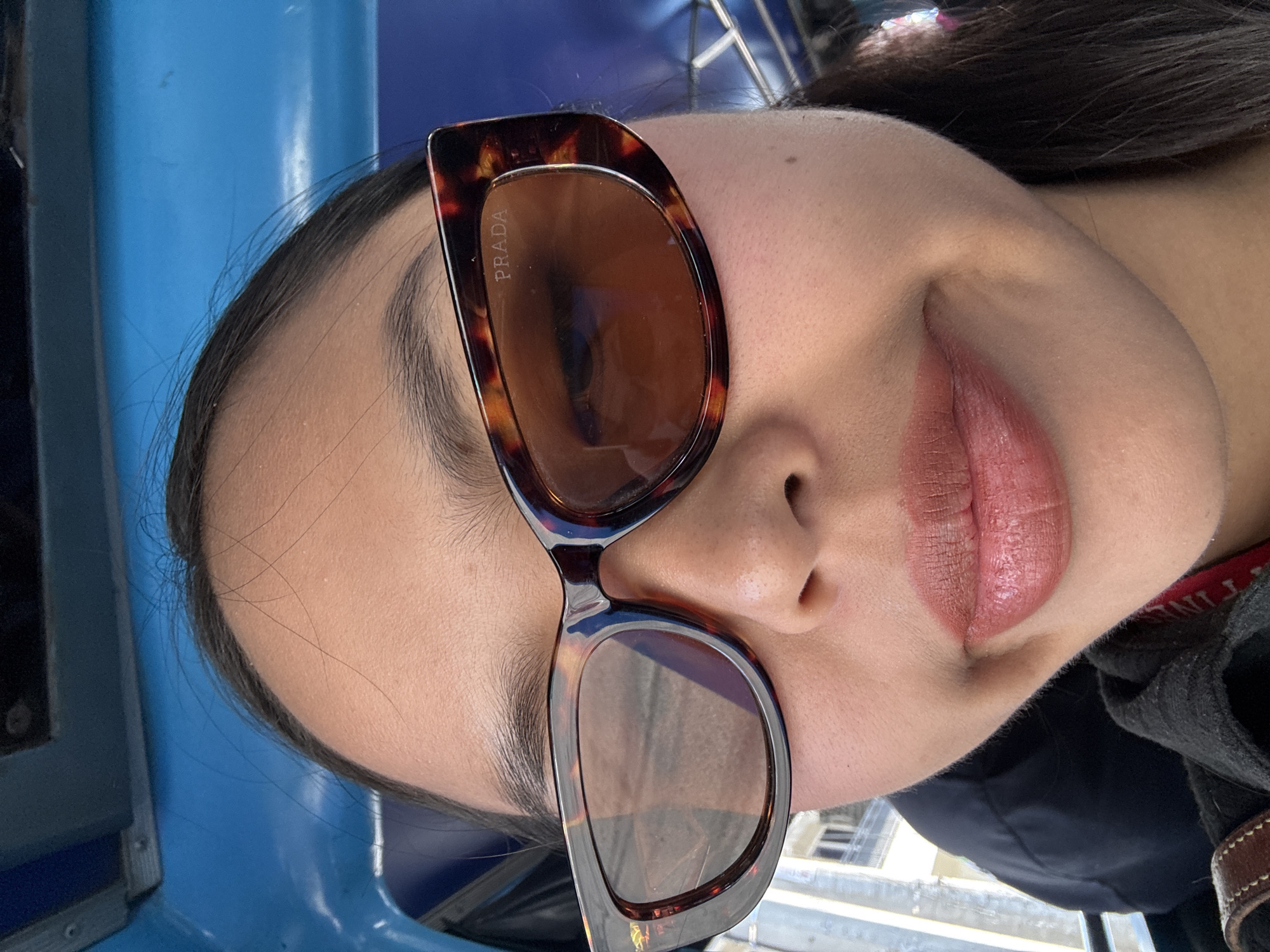 A close-up, canted, and primarily frontal view depicts a woman wearing Prada sunglasses inside what appears to be a public transportation vehicle. The woman has dark hair pulled back from her forehead, and her lips are coated in a neutral-toned lipstick. The sunglasses have a tortoise shell frame and are angled in a way that obscures parts of her face. Reflected in the lenses are blurred images of what seems to be a cityscape. The blue color of the vehicle's interior is visible behind her head. The environment appears urban. No explicit indications identify the location as Davao City, Philippines, although the presence of a public transport vehicle common in the Philippines is an indicator.