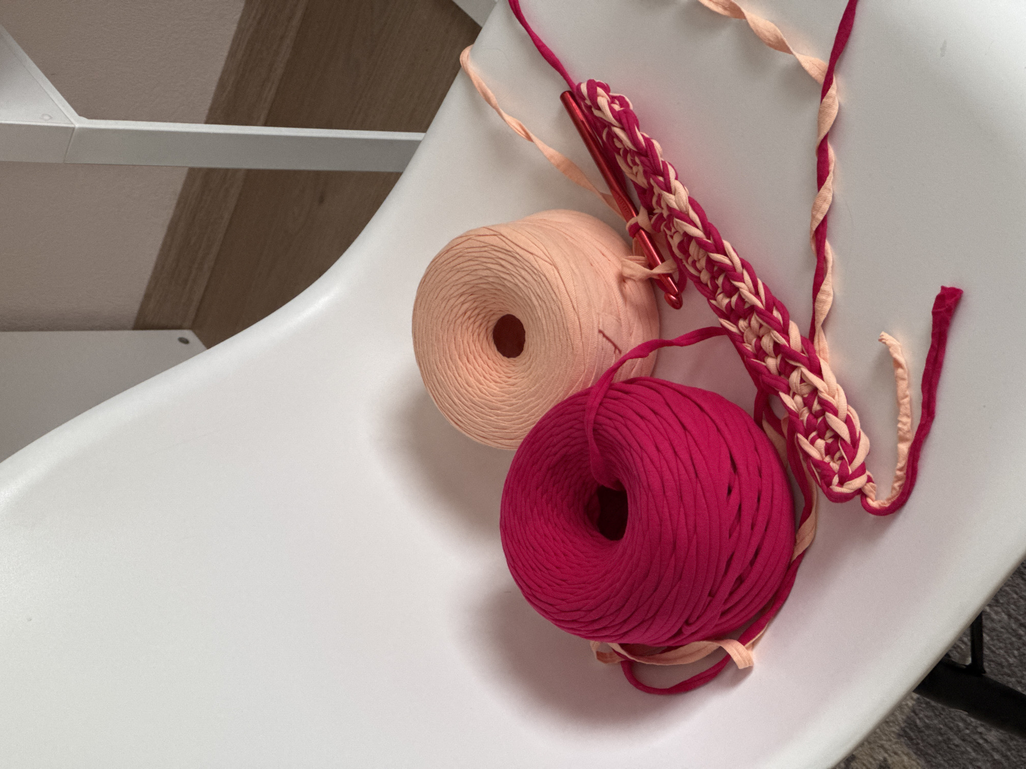 This image captures a close-up, slightly elevated view of crafting materials, likely an ongoing or paused crochet project, set indoors in Izhevsk, Russia. The items are neatly arranged on a white, possibly plastic, chair or furniture surface.
The primary subjects are two large rolls of t-shirt yarn. One roll is a soft peach or light orange color, and the other is a vibrant fuchsia or deep pink. Next to these yarn rolls, two metallic red crochet hooks are visible, embedded in a partially completed piece of crochet work. This piece is a chain of stitches, demonstrating an alternating pattern using both the peach and fuchsia yarns, creating a striped or braided effect. Loose strands of both colored yarns are also present, with some sections twisted together.
In the background, elements of an indoor setting are visible, including a white, angular shelf or furniture frame and a light brown wooden surface, possibly part of a floor or wall paneling. The lighting appears bright and natural, casting soft shadows. There are no people visible in the image, nor is there any discernible text. The scene conveys a sense of creative activity and preparation for a craft project.