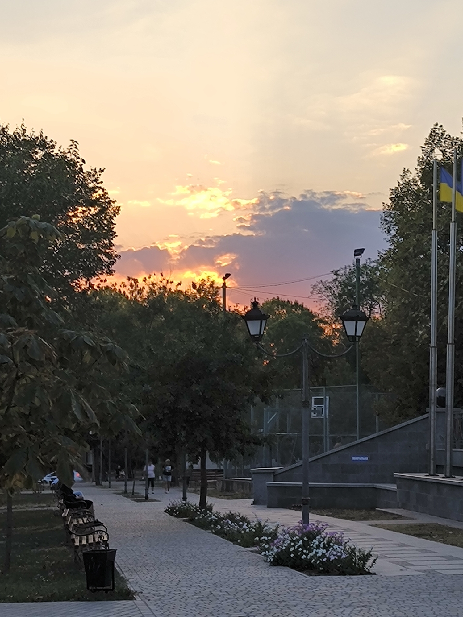 The image captures a sunset scene in a public park or promenade in Mykolaiv, Ukraine. The sky is filled with warm hues of orange, yellow, and pink, with scattered clouds tinged by the setting sun. In the foreground, a paved walkway lined with benches and trees leads into the distance. Several people are visible walking along the path, appearing to enjoy the evening. On the right side of the image, two flags, one of which is the Ukrainian flag, fly from tall poles, suggesting a civic or memorial area. Streetlights are positioned along the walkway, hinting at the transition from day to night. In the background, a fenced area with what appears to be a basketball court is visible, surrounded by trees. The overall atmosphere is peaceful and serene.