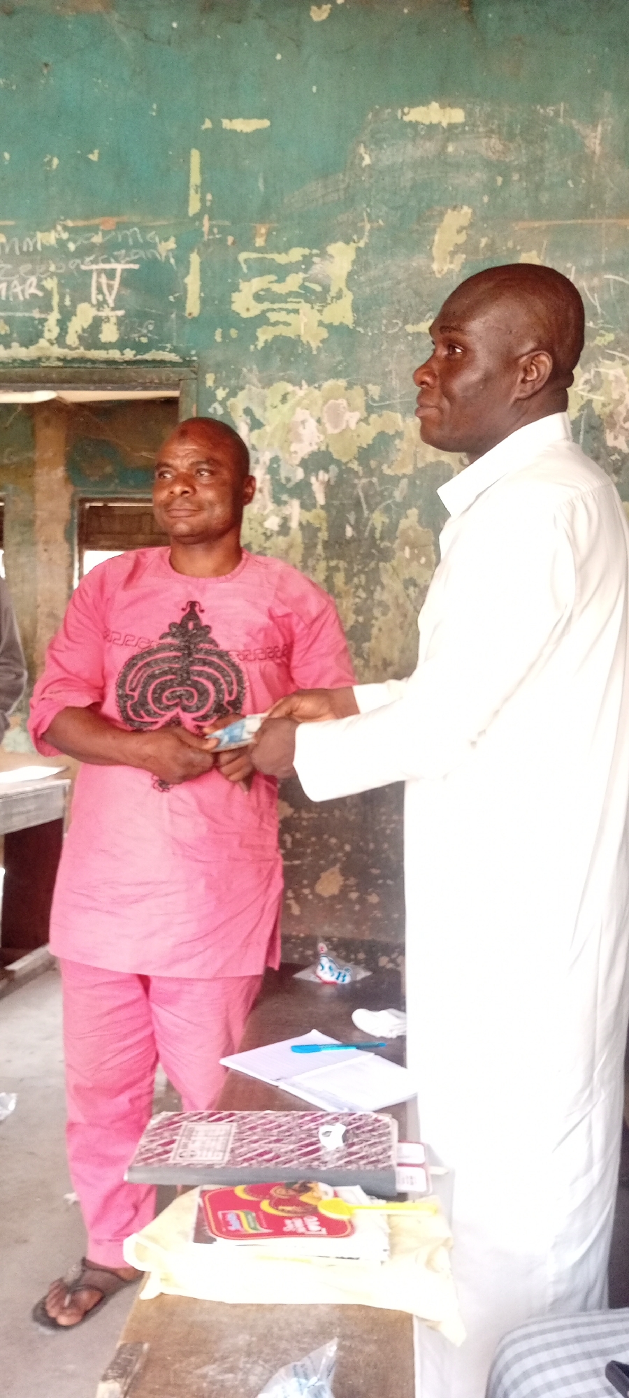 The image captures two men in an indoor setting, possibly a public office or community space, located in an urban area. The first man, on the left, wears a pink traditional outfit and is holding a stack of bills in his hands. The second man, on the right, is wearing a white long-sleeved shirt and appears to be handing over or receiving money. A table in the foreground holds various objects, including notebooks, a pen, and some loose papers. The wall in the background is a faded green color with remnants of old writings and markings.