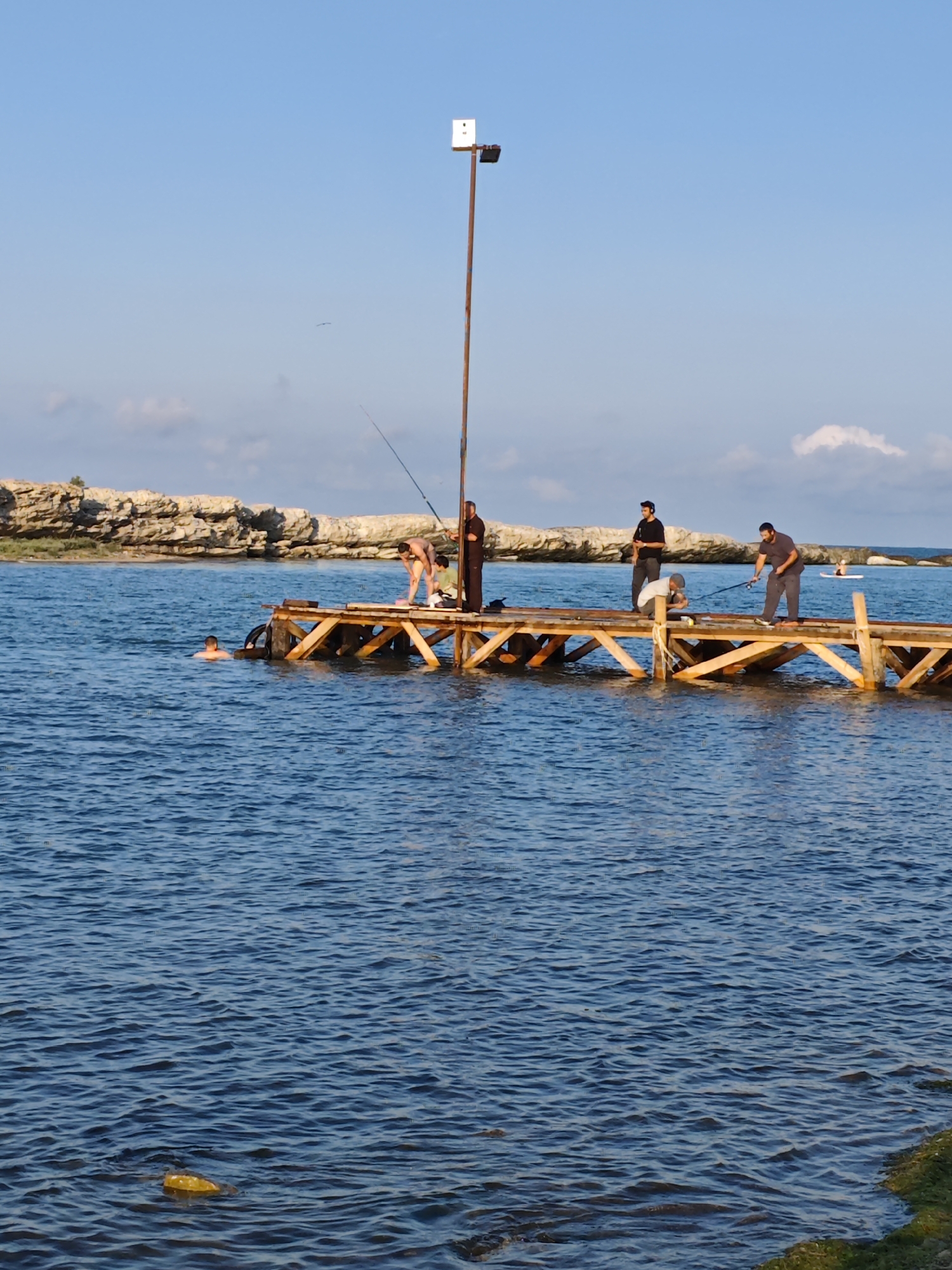 A group of men are fishing from a wooden pier on a sunny day in Izberbash, Russia. The pier extends into a body of calm, blue water, with a rocky shoreline visible in the background. One man is swimming in the water near the pier. The sky is clear blue with a few wispy clouds. A tall pole with a light fixture stands on the pier, and fishing rods are extended over the water. Several men are engaged in fishing, with some casting lines and others tending to their equipment. The overall scene depicts a leisure activity taking place in a coastal environment.