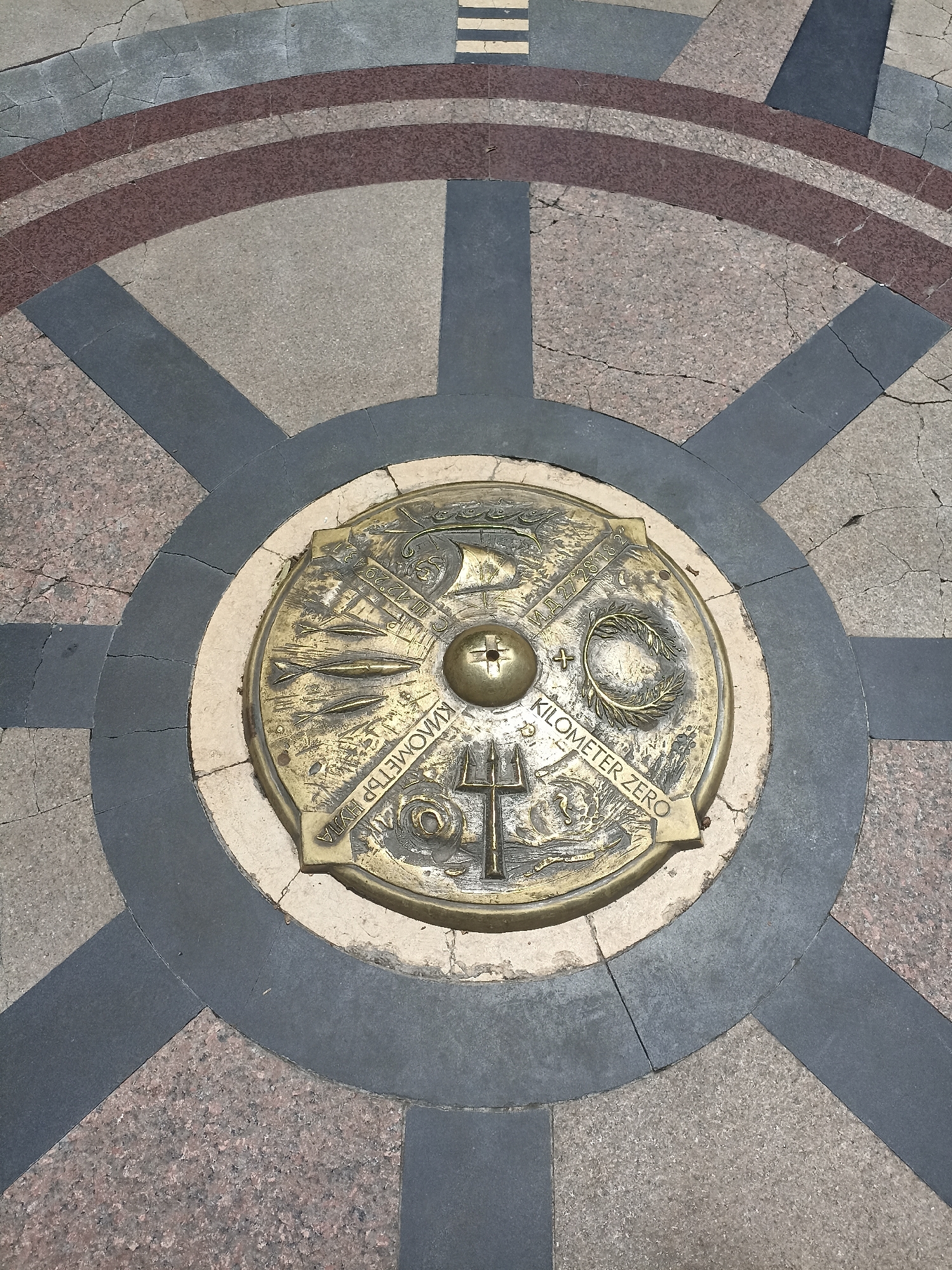 An overhead view captures a bronze "Kilometer Zero" marker, centrally embedded in a circular pattern of paving stones, likely in a public square in Burgas, Bulgaria. The main subject is a circular bronze disk, appearing aged and tarnished, featuring intricate bas-relief designs.
At the very center of the bronze disk is a raised spherical element with an incised cross. The disk is divided into quadrants, each containing distinct symbols. Visible imagery includes a sailing ship, a partial compass rose, a laurel or olive wreath, a group of fish or dolphins, and a trident.
Prominently inscribed on the marker are the words "KILOMETER ZERO" in English and "КИЛОМЕТЪР НУЛА" in Bulgarian, unequivocally stating its function as a central geographical reference point. Also present are coordinate markings: "С.Ш. 42.29.43" (indicating North Latitude 42 degrees, 29 minutes, 43 seconds) and "И.Д. 27.28.18" (indicating East Longitude 27 degrees, 28 minutes, 18 seconds).
The bronze marker is framed by a lighter, off-white circular band of stone, which is then surrounded by a broader, decorative pattern of paving. This surrounding pavement consists of dark grey, light grey, and reddish-brown granite tiles arranged in radiating spokes and concentric arcs, reminiscent of a large compass rose or a ship's wheel, complementing the maritime and navigational themes of the central disk. Several cracks are visible in the surrounding pavement, suggesting wear and the passage of time.
The scene is an outdoor setting during daytime, with soft, even lighting and no harsh shadows. No people or specific activities are depicted, focusing entirely on the static, symbolic marker. The overall impression is one of historical and geographical significance within an urban landscape.