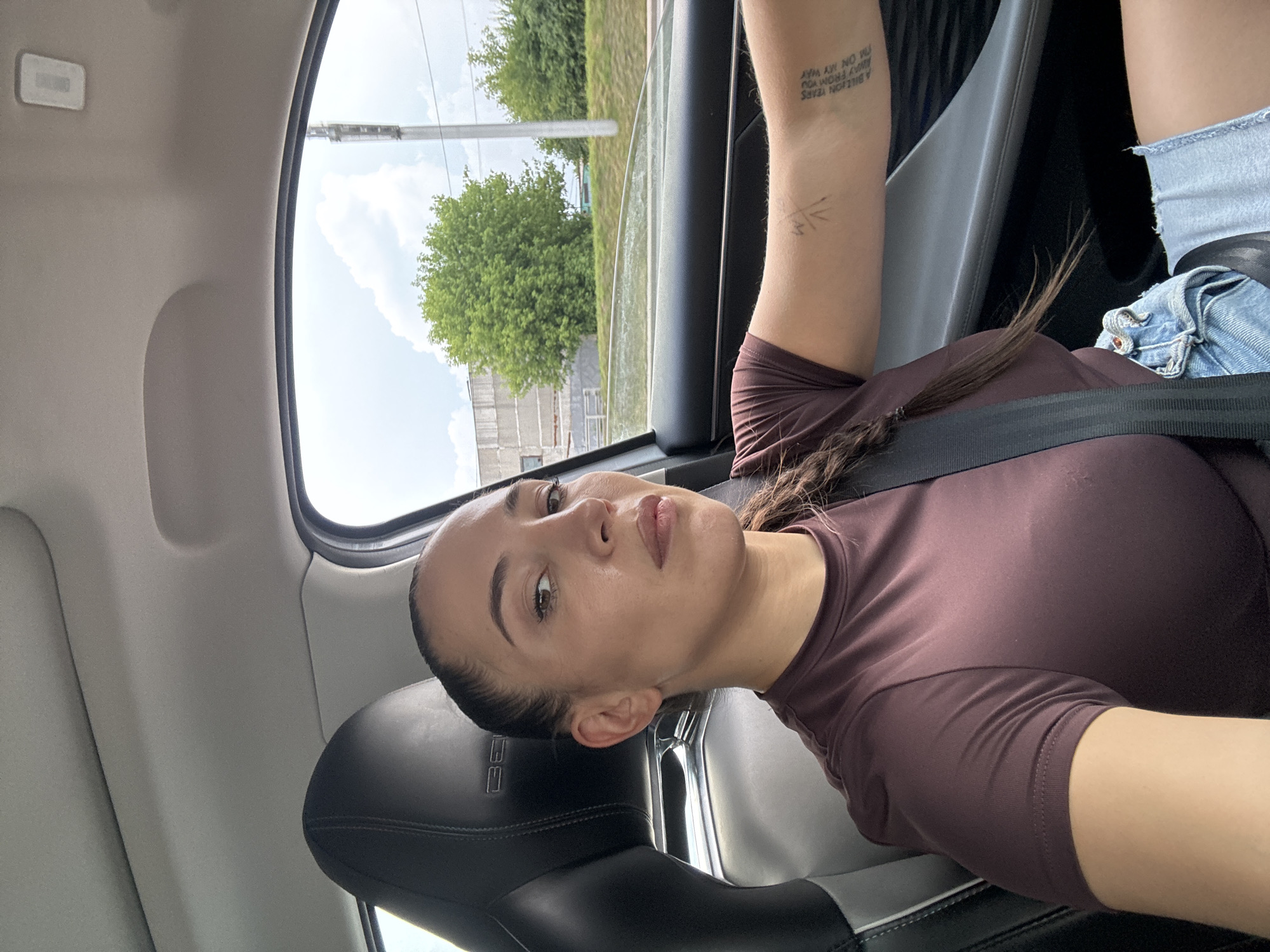 A woman is sitting in the driver's seat of a car, looking at the camera. She has her hair in a braid and is wearing a maroon t-shirt and denim shorts. She has a tattoo on her left arm. The car appears to be in motion, as indicated by the scenery outside the window, which includes trees and buildings. The scene takes place in the city of Krasnaya Pakhra, Russia.