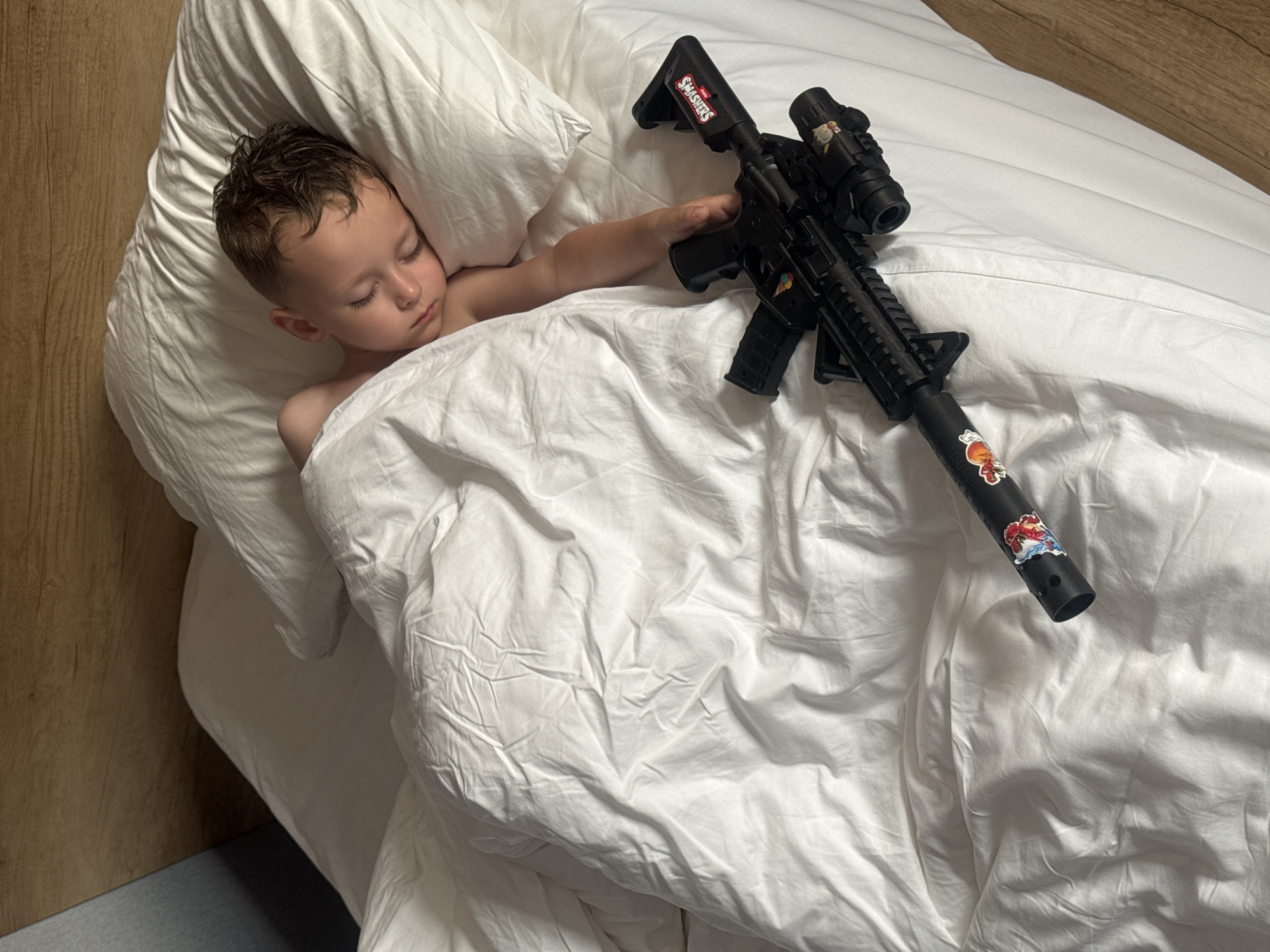 A young boy, with dark hair and closed eyes, sleeps soundly in a bed covered with white sheets. His head rests on a white pillow. To the right of the boy, partially visible under the sheets, is a black toy rifle. The rifle has stickers on it, including one with the word "SMASHERS" on the stock and another with an image of what appears to be a cartoon character. The boy's right arm is extended, and his hand rests on the toy rifle. The scene appears to be indoors, possibly in a bedroom, with a wooden headboard visible in the background. The lighting suggests it is daytime. There are no recognizable landmarks or indicators of the city of Qinhuangdao, China in the image.
