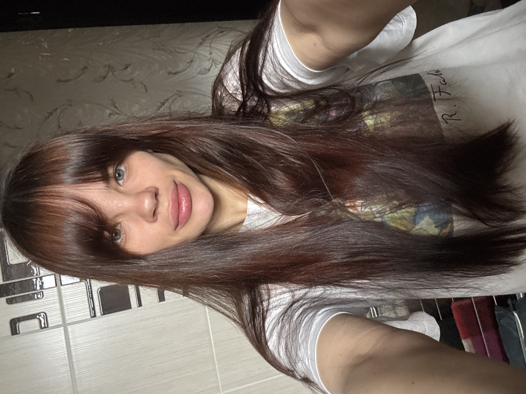 A woman with long, brown hair and bangs is posing for a selfie. She has light skin and blue eyes. She is wearing a white t-shirt with an image and the signature "R. Fah". The background includes a wall with a decorative pattern and a tiled surface with black accents. The location is possibly in an apartment or house, but there is no information to confirm it is in Rostov-on-Don, Russia.