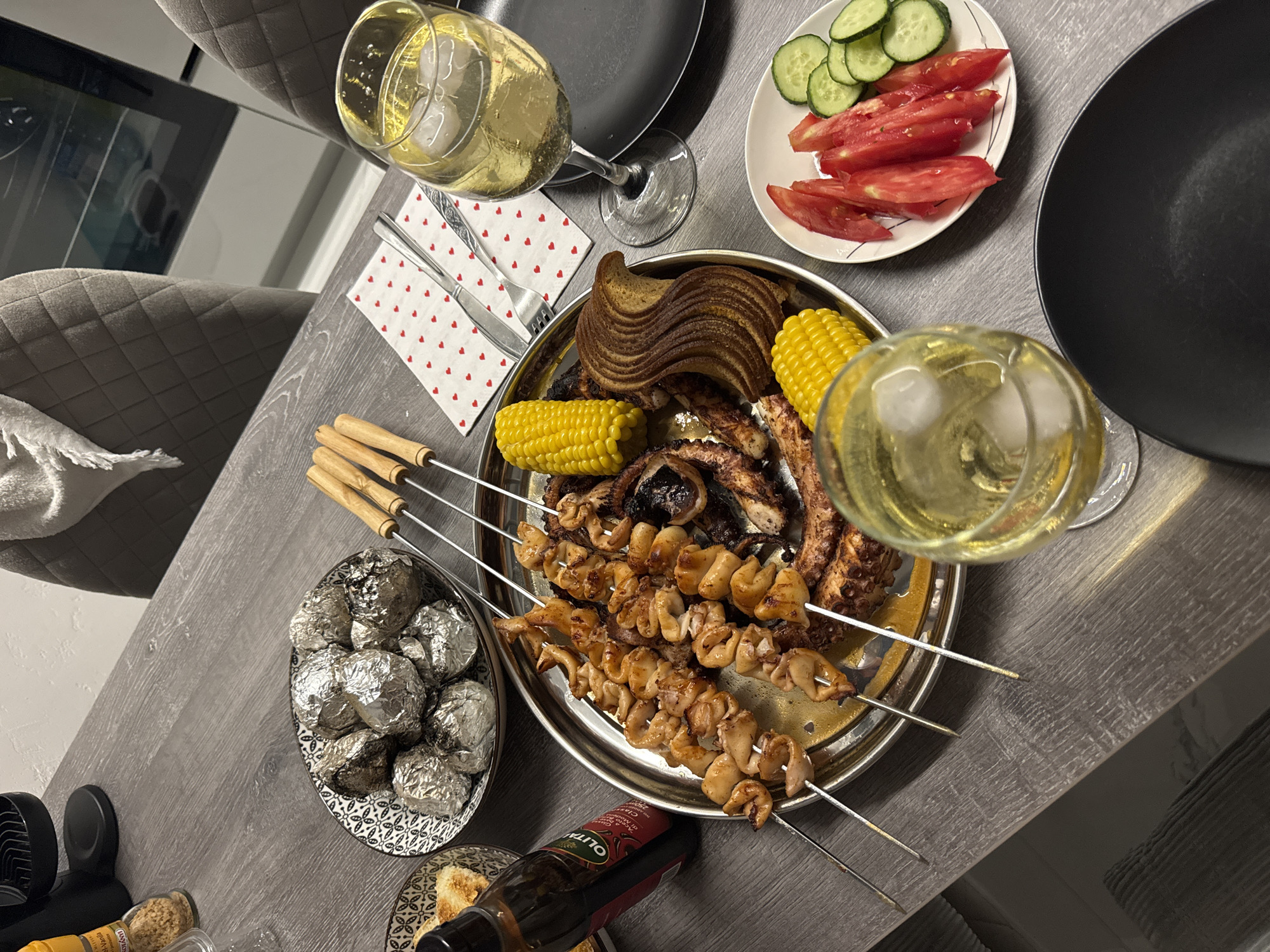 An overhead, medium shot captures a dinner spread on a wooden table. In the foreground, two glasses of white wine with ice sit beside black plates. A white plate in the middle right holds sliced tomatoes and cucumbers. A large silver platter in the center showcases grilled octopus skewers, corn on the cob, and a loaf of sliced bread. To the left, a bowl contains foil-wrapped potatoes and a bottle of olive oil is visible. The scene is set in a dining room, likely in the city of Sustaš, Montenegro, judging by the context. There are no people visible in the image.