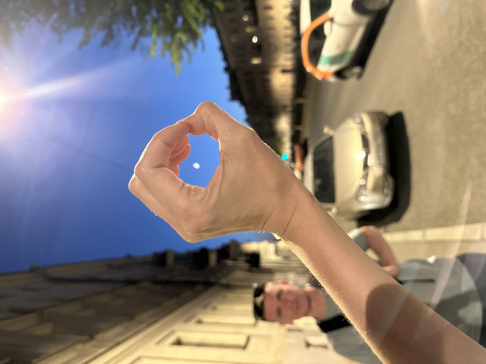 A person is making an "OK" hand gesture in an outdoor setting, likely Saint Petersburg, Russia, based on architectural style. The background features a street with parked cars, buildings, and street lighting at dusk or night. The person's face is visible, along with a portion of their arm and clothing. The sky is transitioning from blue to dusk.