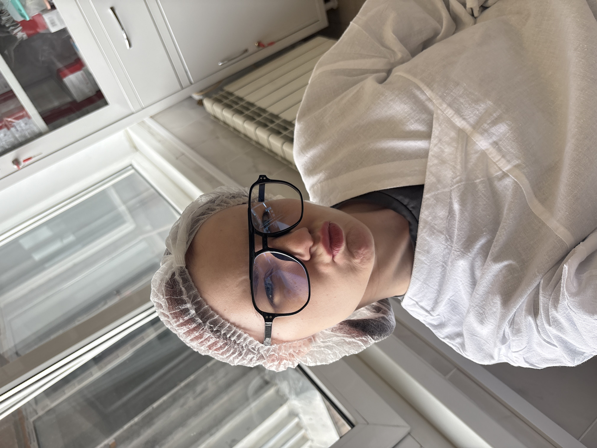A young woman wearing a disposable hair net and oversized black-rimmed glasses poses for a selfie. She is wearing a white lab coat over a dark t-shirt. Her lips are pursed in a duckface. The background shows a window with a reflection of what appears to be a city skyline, a white cabinet with drawers, and a radiator. The overall environment suggests a clean, possibly medical or laboratory setting, within a building in an urban area. The lighting is bright, coming from the window.