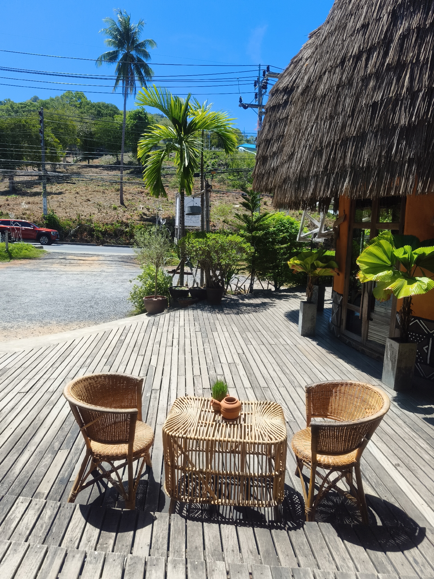 This is a high-angle, sunlit outdoor photograph taken at what appears to be a resort or cafe in Baan Thong Po, Thailand. In the foreground, a wooden deck is furnished with two woven rattan chairs and a small, similarly constructed table. The table holds two small terra cotta pots, one with a sprig of greenery. The deck planks are weathered and laid horizontally. Beyond the deck, a gravel area and a road are visible, with a red vehicle parked on the side. A lush, green hillside with trees, including a tall coconut palm and a smaller fan palm, rises in the background. Overhead, power lines crisscross the bright blue sky. To the right, a building with an orange exterior features a thatched roof and large windows. The overall scene suggests a tropical, relaxed atmosphere.