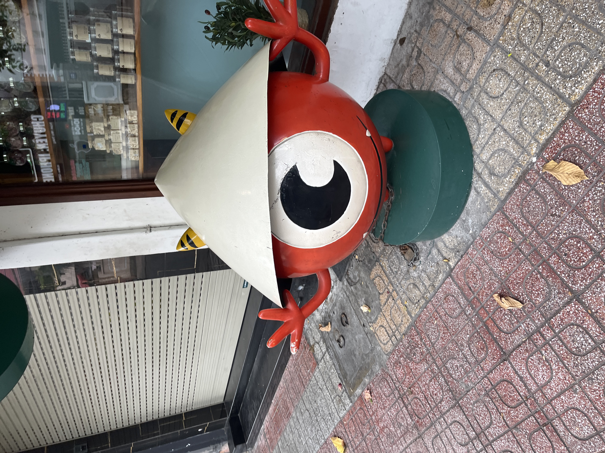 A quirky, red, cartoon-like creature with large white eyes and black pupils is positioned on a green circular base on a tiled sidewalk in front of a shop. The creature is wearing a conical, pale yellow hat that casts a shadow over its face, and has two yellow and black striped horns peeking out from under the hat. Its red hands are extended outwards, with one hand raised as if waving. The shop behind the creature has a large window displaying various jars and products on shelves, and a metal roll-up shutter. The sidewalk is paved with red and gray square tiles, some of which are decorated with a swirling pattern. A few dry leaves are scattered on the ground. The scene appears to be outdoors on a street in Nha Trang, Vietnam.