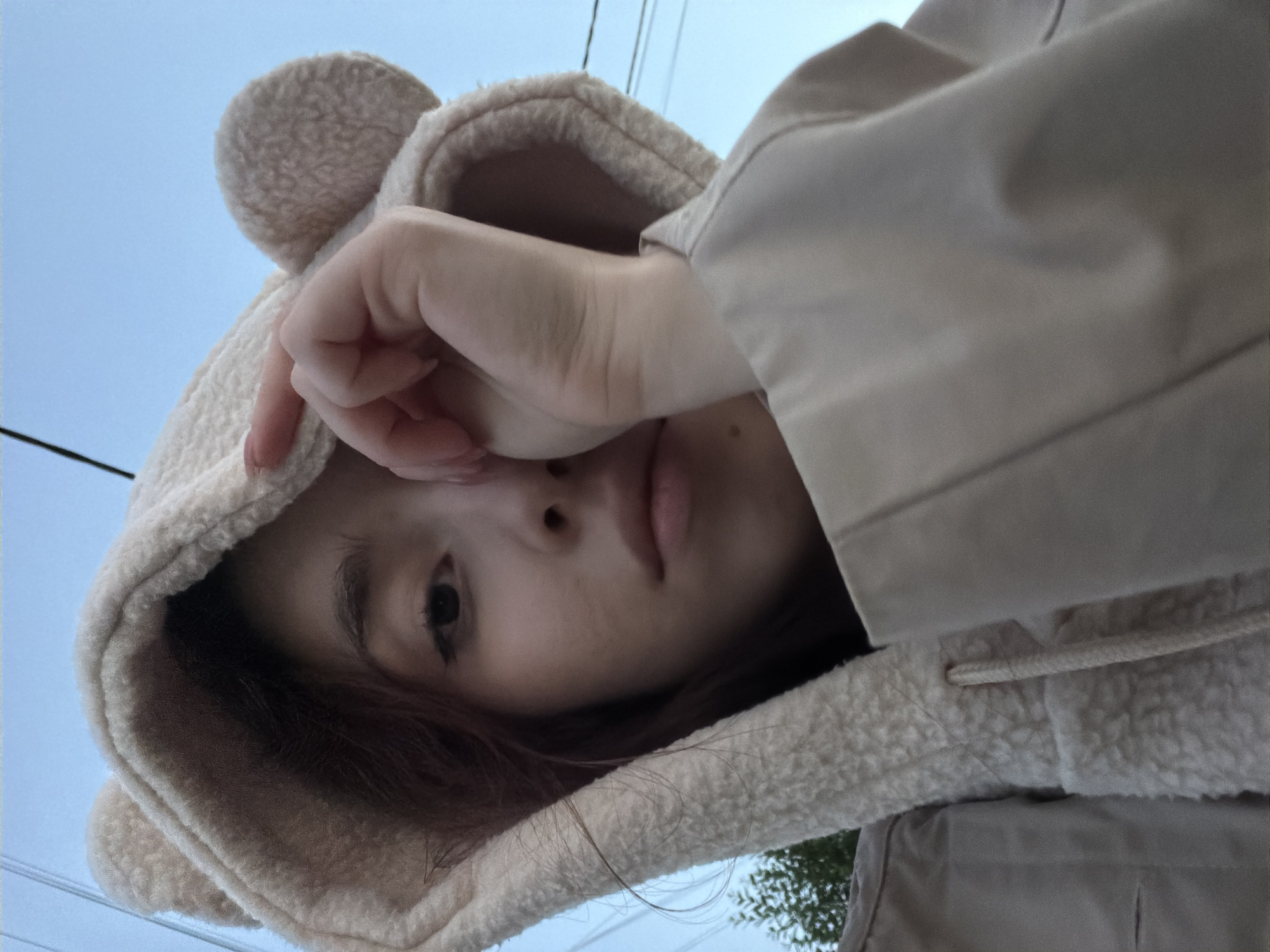 A young woman is captured in a close-up, low-angle selfie. She is wearing a beige teddy bear-eared hoodie and a beige jacket. Her right hand is raised, partially obscuring her right eye and forehead. The background consists of a clear blue sky with visible utility wires. To the left, a small portion of a green tree is visible. The lighting suggests it is either dawn or dusk. The woman has dark hair and appears to be outdoors. The overall setting is urban, but no specific landmarks are identifiable.