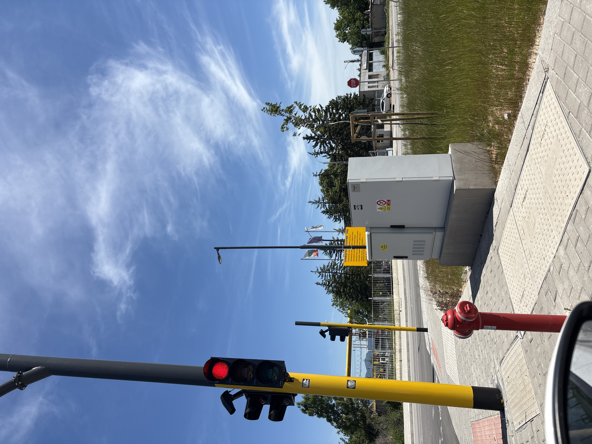 The image shows a street scene in an unknown city. The camera is angled upward, capturing a traffic light displaying a red signal. The traffic light is mounted on a yellow pole and is partially obstructed by the side mirror of a vehicle. To the right of the traffic light, a red fire hydrant is visible on a paved sidewalk. Behind the hydrant, there is a large grey electrical box with warning signs posted on it. In the background, several flags are flying from a tall pole, and a stop sign is visible near a grassy area and buildings. The sky is blue with wispy white clouds.