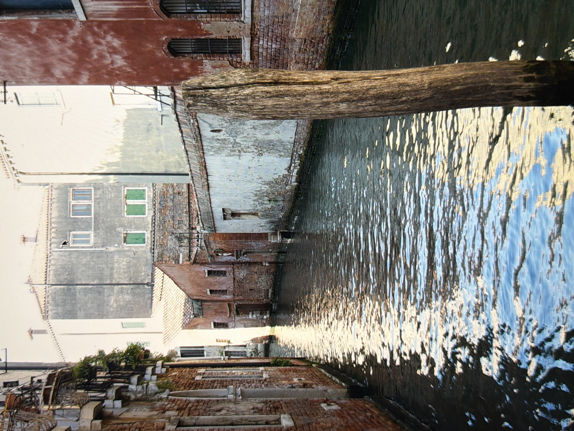 This is a photograph of a canal in Venice, Italy. The canal is lined with old brick buildings. The water reflects the sunlight, creating a shimmering effect. A weathered wooden post stands in the foreground, to the right of the canal. The scene is tranquil and evokes a sense of history and timelessness.