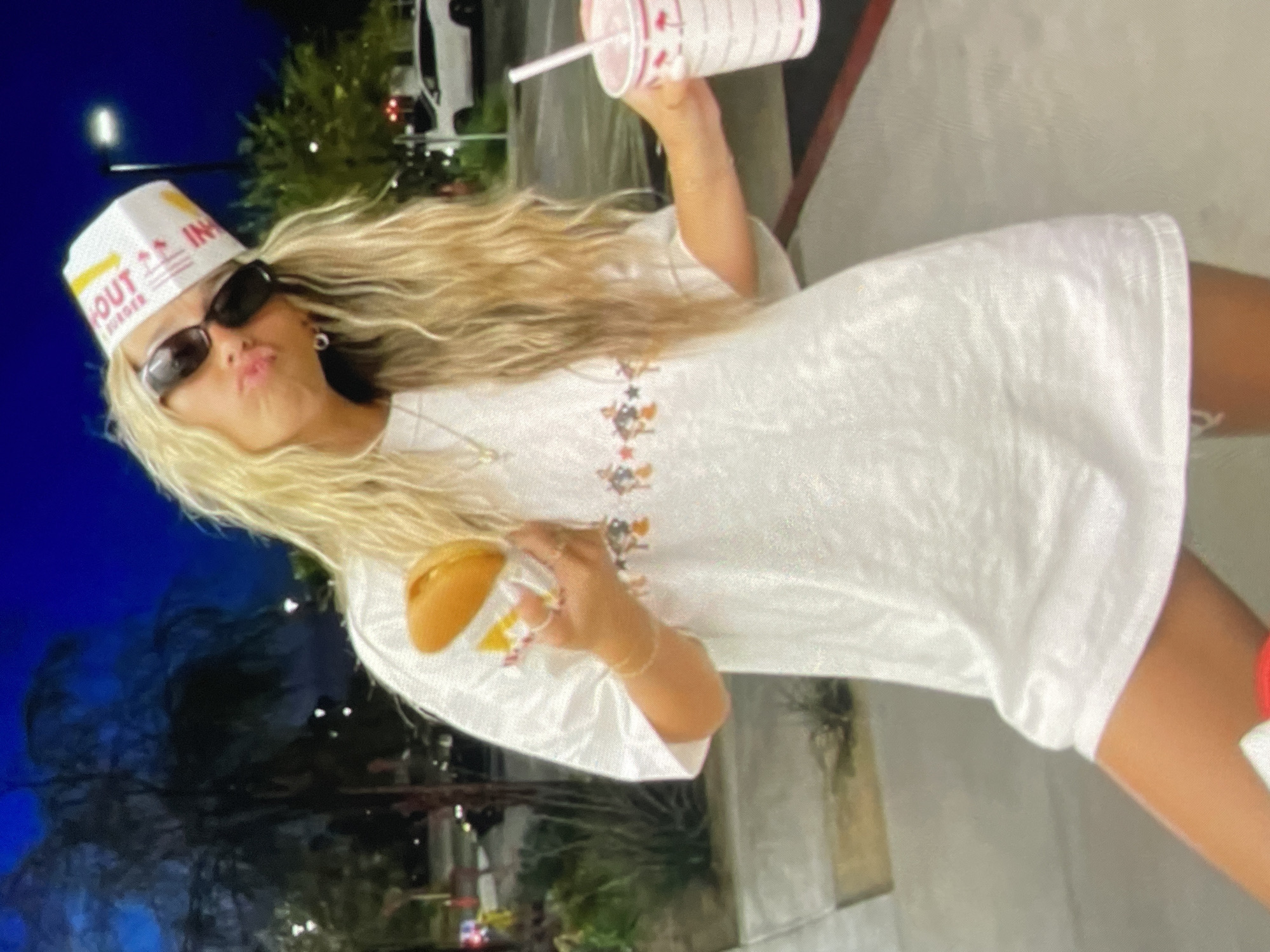 A woman with long blonde hair, sunglasses, and an In-N-Out Burger hat is holding a burger in one hand and an In-N-Out Burger drink in the other. She is wearing a white t-shirt. The location is indeterminate, but NOT Harmanli, Bulgaria.