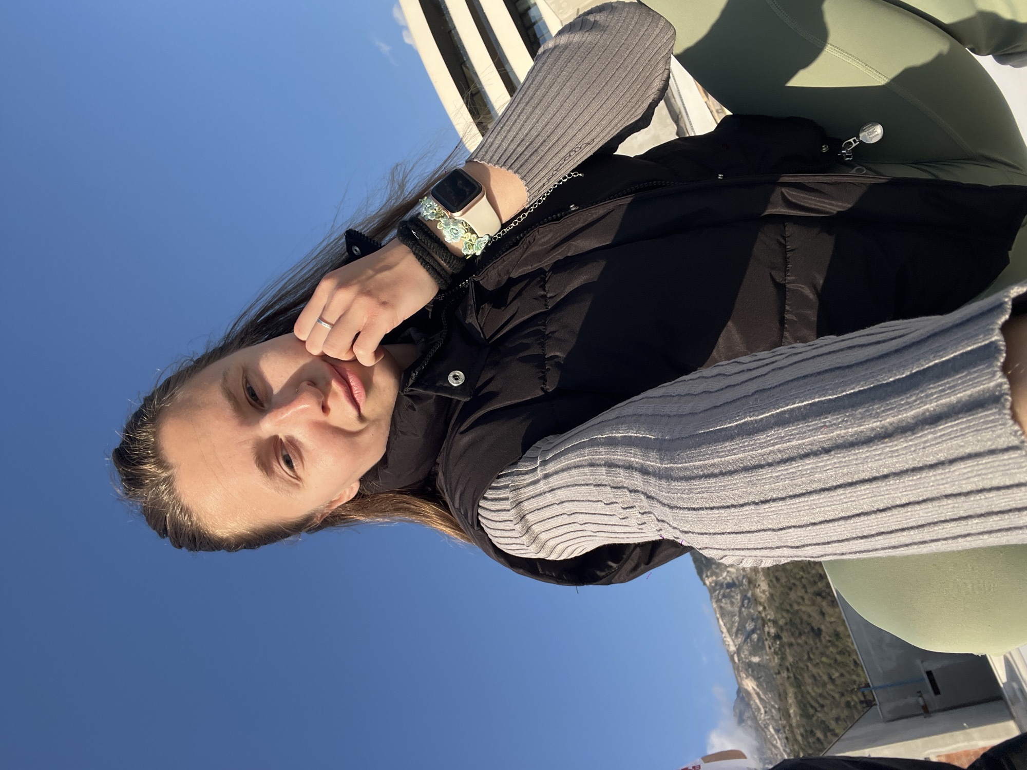 The image shows a woman in a grey sweater and black vest. She has brown hair and is wearing a watch and a ring. The background includes a blue sky, a building or structure with vertical elements, a mountainous area, and what appears to be green clothing. Based on the limited background information, the location might be in or near Rafailovići, Montenegro, given the presence of mountains and coastal structures.