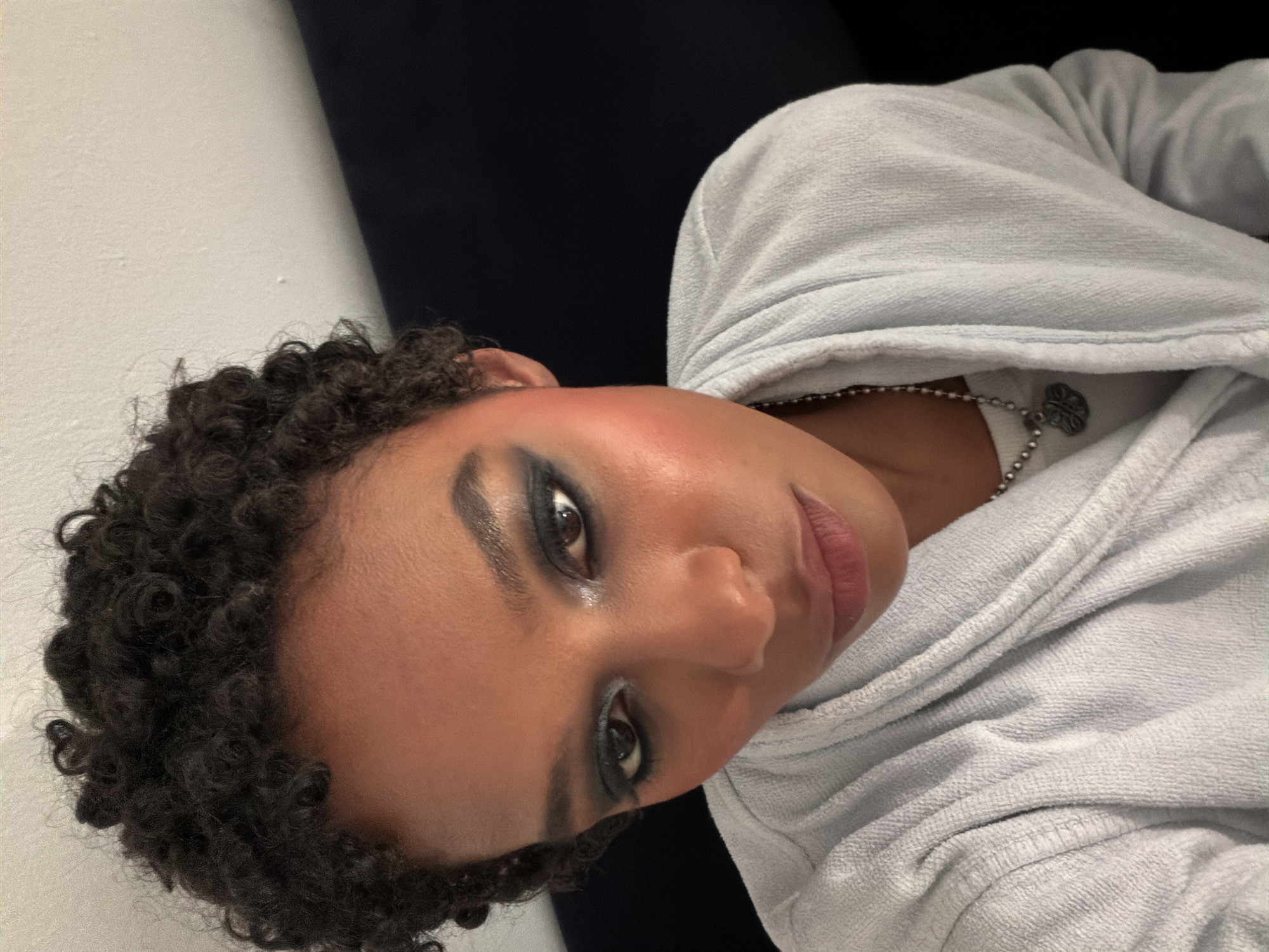A person with dark, curly hair and smokey eye makeup is seen indoors. The person is wearing a light blue hoodie. A silver necklace with a charm is visible. The background consists of a white wall and a dark blue or black surface. No evidence suggests the photo was taken in Bagnolet, France.
