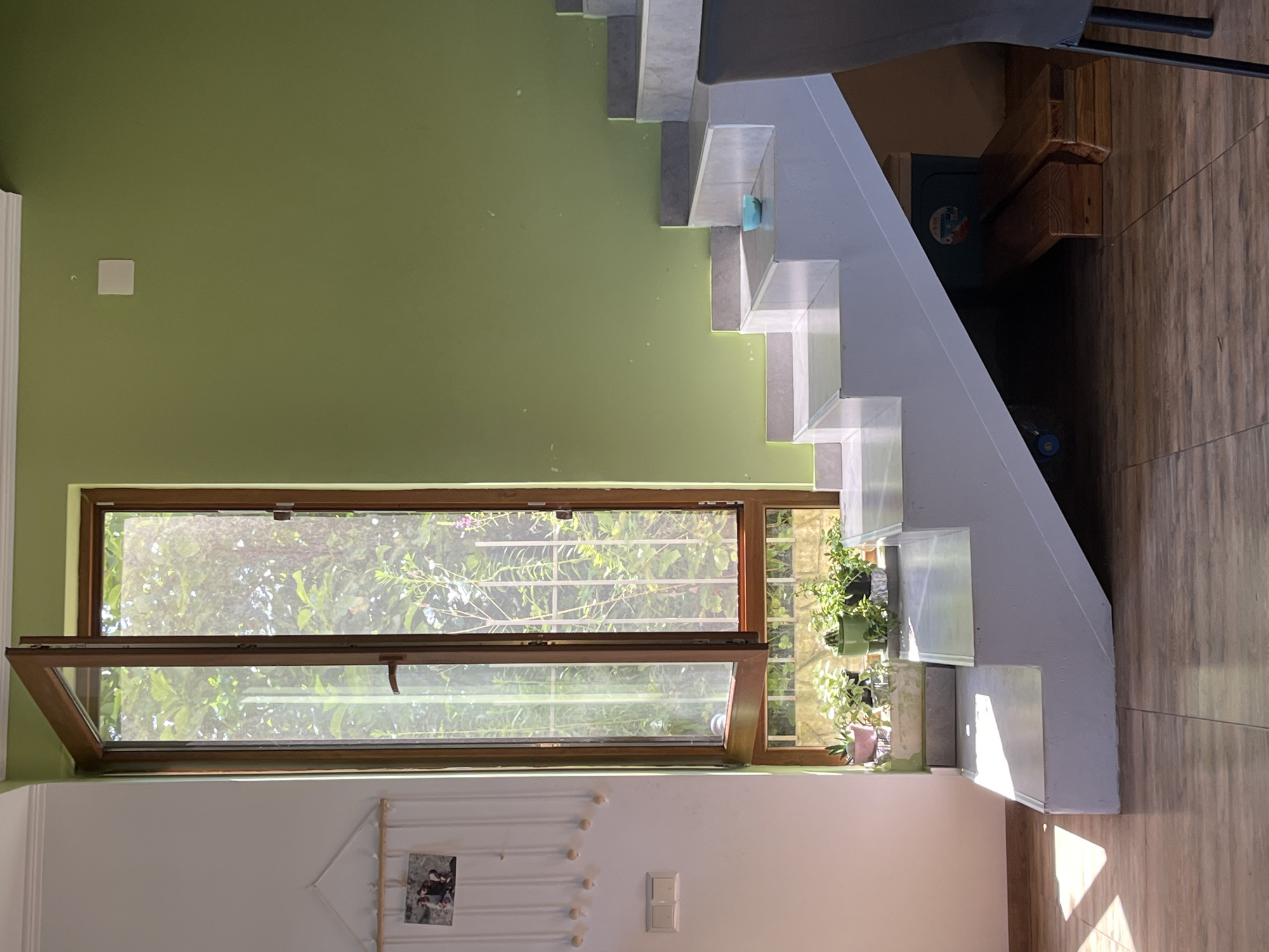 The image depicts an interior scene, possibly a residential space, with a prominent staircase and an open window. The staircase is modern and white, with what appears to be grey marble trim. Sunlight streams in from the open window, illuminating the lower steps of the staircase and casting geometric shadows on the tiled floor.
On a ledge near the bottom of the window, several potted plants are visible, including what looks like a cactus and other greenery. The window itself is framed by wood and looks out onto a garden or outdoor space with lush foliage.
To the left of the window, a white wall features a decorative tapestry with strings and small wooden balls, and a picture frame holding two photographs. Below this, a light switch with two toggles is installed on the wall. The wall to the right of the staircase is painted a vibrant, light green.
Underneath the staircase, there is a dark space where two wooden planks are placed side-by-side, possibly forming a makeshift bench or support. A blue cooler or container with a label is also partially visible in this area. A dark grey chair with black legs is partially visible in the lower right corner of the frame, suggesting a dining or seating area.
The floor is covered in light brown tiles with a wood-grain pattern. The overall impression is of a bright, airy space with a mix of modern and natural elements. This scene is located in the city of Ureki, Georgia.