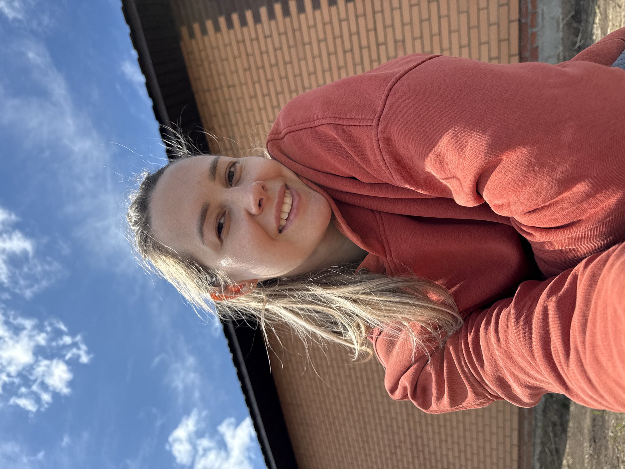 A young woman with blonde hair is smiling at the camera. She is wearing a red-orange hoodie. Behind her is a brick building and a blue sky with clouds. The location is Chaikovsky, Russia.