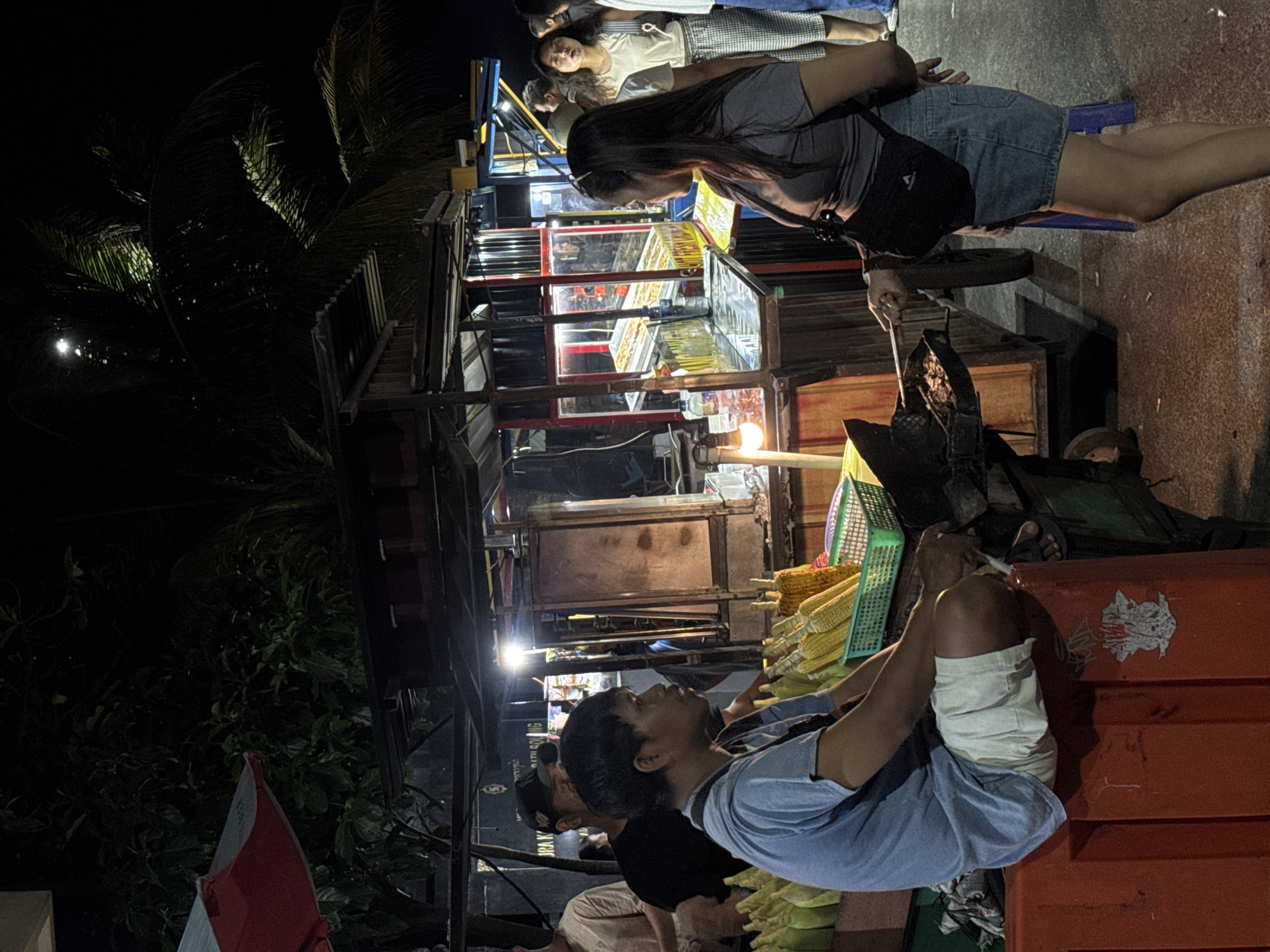 The image depicts a night scene at a street food market in Canggu, Indonesia. A woman with long dark hair, wearing a gray shirt and denim shorts, sits on a small blue stool, using a long metal utensil to stir something in a wok. Several people stand behind her. In the foreground, a man wearing a light blue t-shirt is sitting and smoking a cigarette. He is surrounded by ears of corn. The background features food stalls brightly lit with neon lights, displaying various items for sale. Tropical foliage is visible in the periphery of the scene.