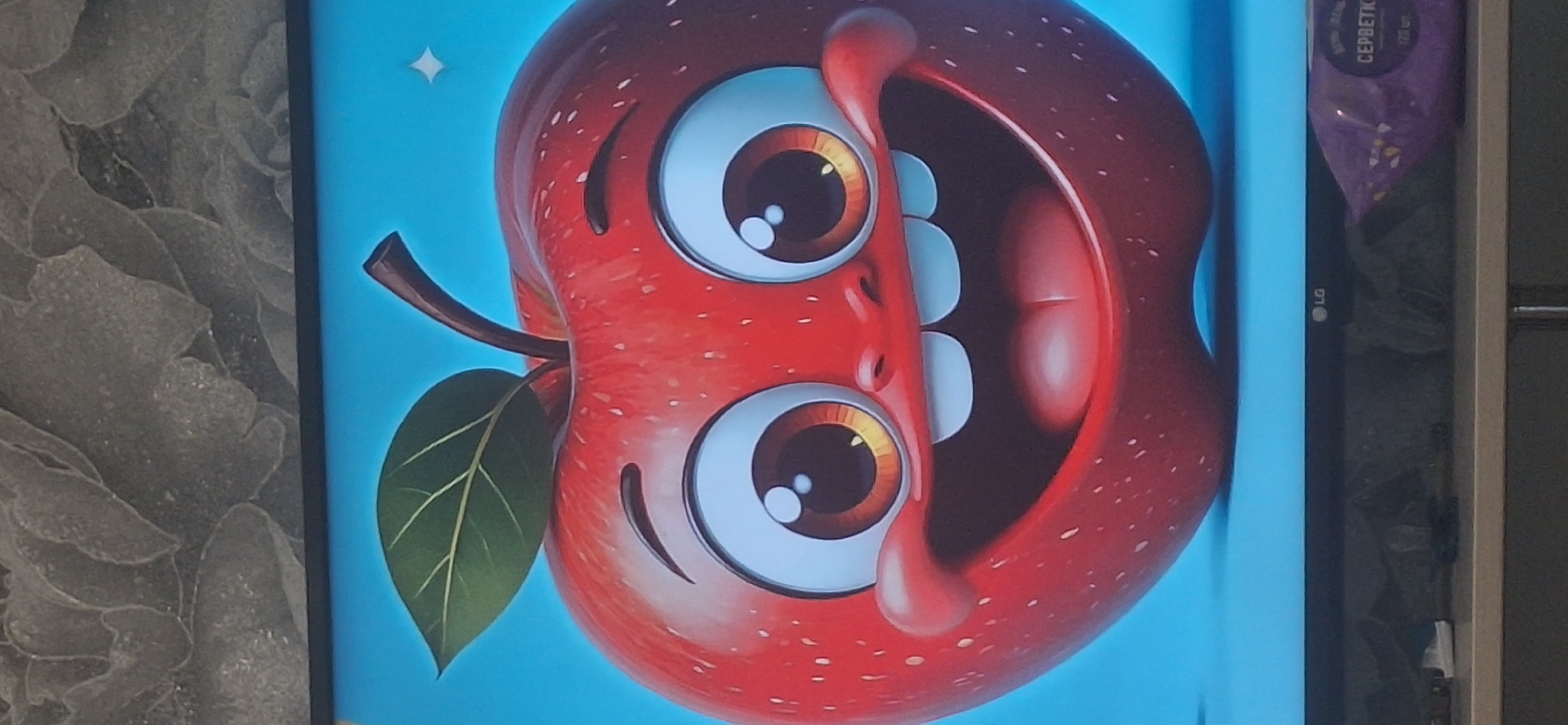 The image displays a close-up of a cartoon apple with a cheerful expression. The apple is bright red with small white speckles and has a green leaf and a brown stem. It features large, wide eyes with orange irises and white pupils, a wide grin showing two small white teeth, and plump, rosy cheeks. The apple is set against a solid blue background. Below the apple, the top edge of a television screen is visible, showing the LG logo. A purple package with Cyrillic text is partially visible on the shelf beneath the television. The scene appears to be indoors, likely a living room or entertainment area. The image itself is a photograph taken of a screen, indicated by the slight glare and the visible edges of the television. The location of Bila Tserkva, Ukraine, is not depicted or implied in this image.
