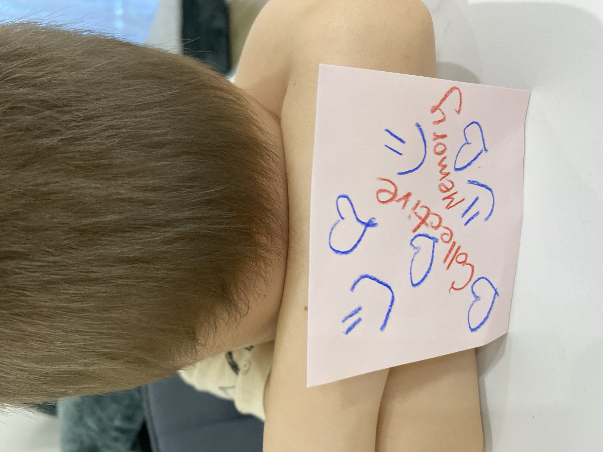A child with short brown hair is holding a pink square of paper that is angled towards the camera. The paper has the words "Collective" and "Memory" written on it in red and blue ink, along with heart symbols. The child's arm is bent, and their head is resting on it. The background is blurry but includes a white surface, a blue-gray object, and a window. The location in the city of Chaikovsky, Russia, is not discernible from the image.