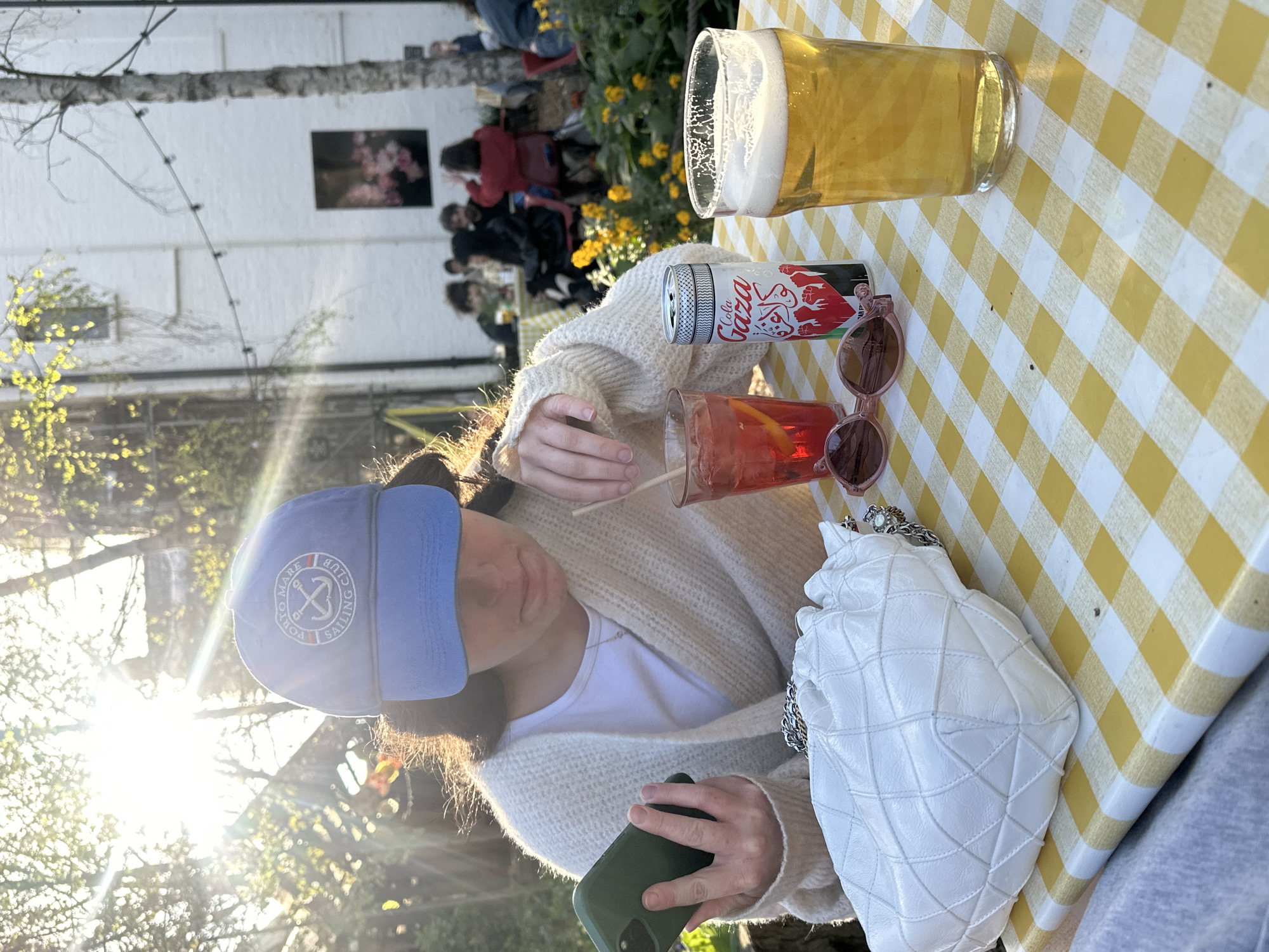 A woman wearing a blue baseball cap, a white T-shirt, and a cream-colored cardigan is seated at a yellow and white checkered table outdoors. She holds a smartphone with a green case in her hands. On the table are a glass of amber liquid with a frothy top, a can of Cola Gaza, a glass of red liquid with a straw, a pair of pink sunglasses, and a white quilted handbag with a silver chain. In the background are several people seated at tables with yellow flowers. A white building with windows and a framed picture is visible in the distance. The setting is illuminated by sunlight. The location is likely London, United Kingdom.