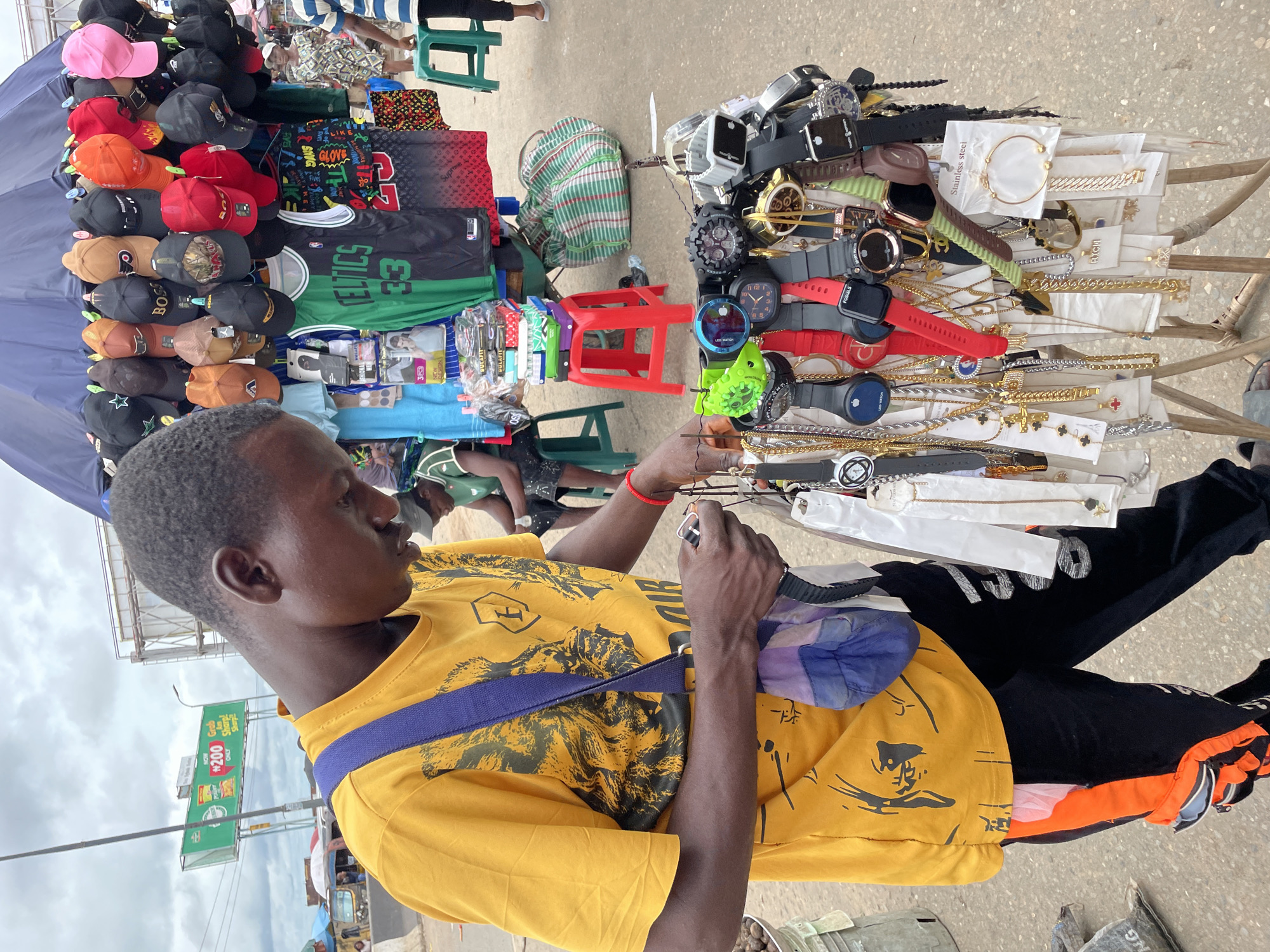 A man wearing a yellow t-shirt with a graphic print and black pants with orange stripes stands beside a display of watches and jewelry. He is looking down at the display, appearing to examine some items. The display is set up on a metal stand with several rows of watches and necklaces. In the background, there are other vendors and shoppers, as well as a large blue umbrella providing shade for a merchandise stall. There are also several baseball caps hanging from the stall. The scene appears to be an outdoor market or street vendor setup in an urban environment.