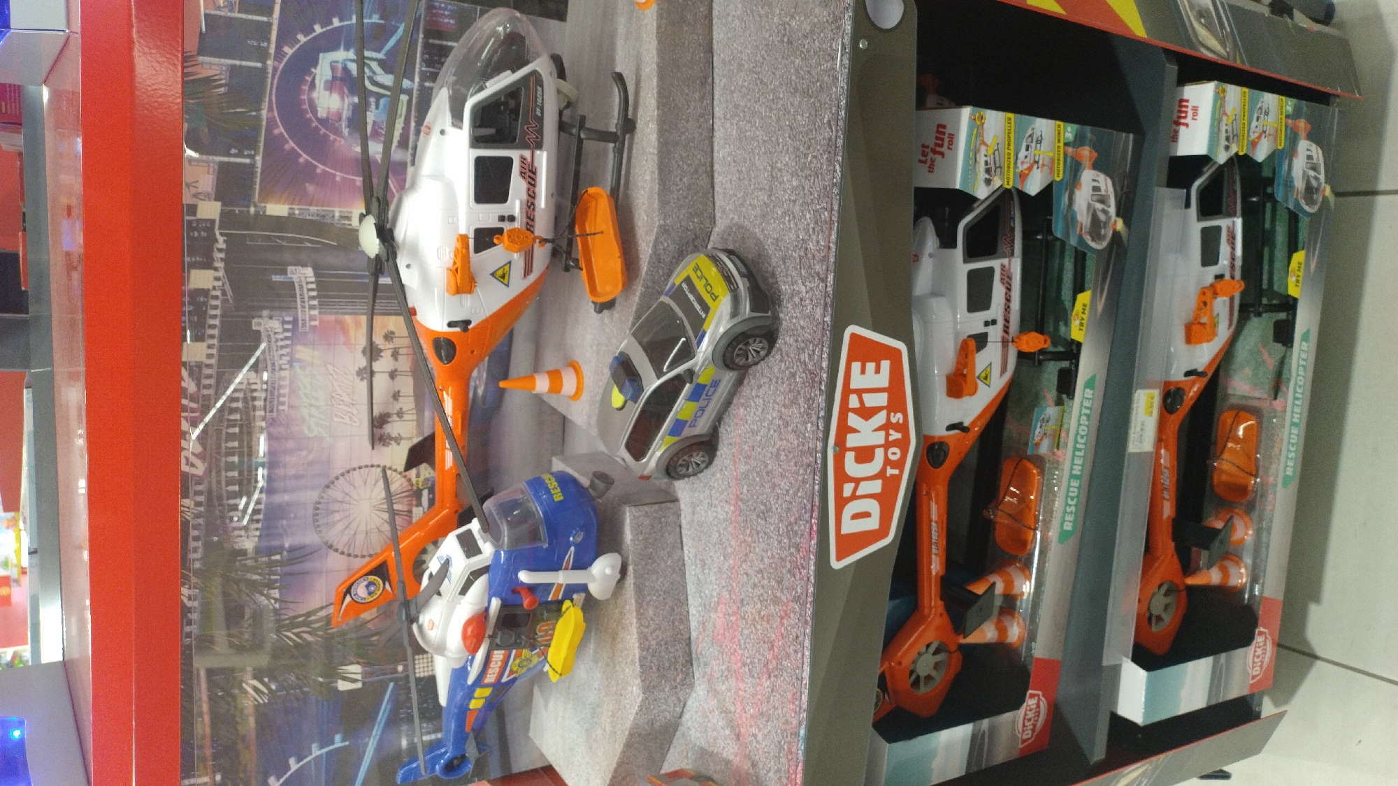 The image shows a retail display of toy rescue vehicles. In the foreground, a display box for "Dickie Toys" rescue helicopters is visible. Two toy rescue helicopters are partially visible inside the box, along with orange traffic cones and a stretcher. Above this box, a diorama features toy rescue vehicles, including a blue and yellow rescue helicopter, a white and orange rescue helicopter with a basket hanging from it, and a silver police car. The backdrop of the diorama is a colorful mural depicting a cityscape with a Ferris wheel and palm trees. The overall scene suggests a toy store or a similar retail environment. The provided crops do not offer additional context about the location within the city of Pylaia Municipal Unit, Greece.