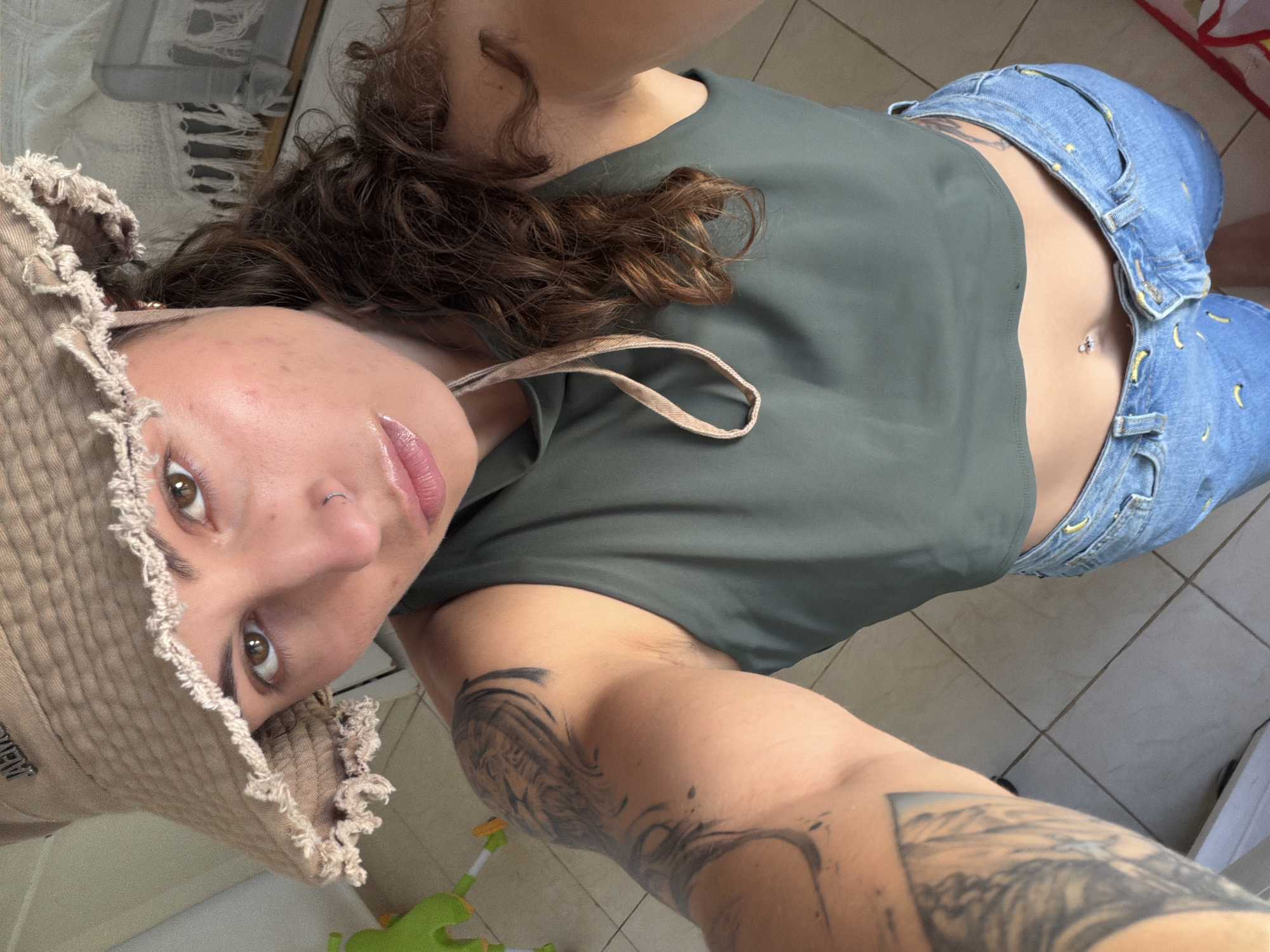 A woman is lying on a tiled floor, taking a selfie from a low angle. She is wearing a green top, blue denim shorts, and a beige bucket hat. She has curly brown hair and visible tattoos on her arms. A section of a white and gray patterned mat is visible in the background along with a green and white child’s toy on the floor near the edge of the frame. There is no indication of the location in Kiryat Bialik, Israel.