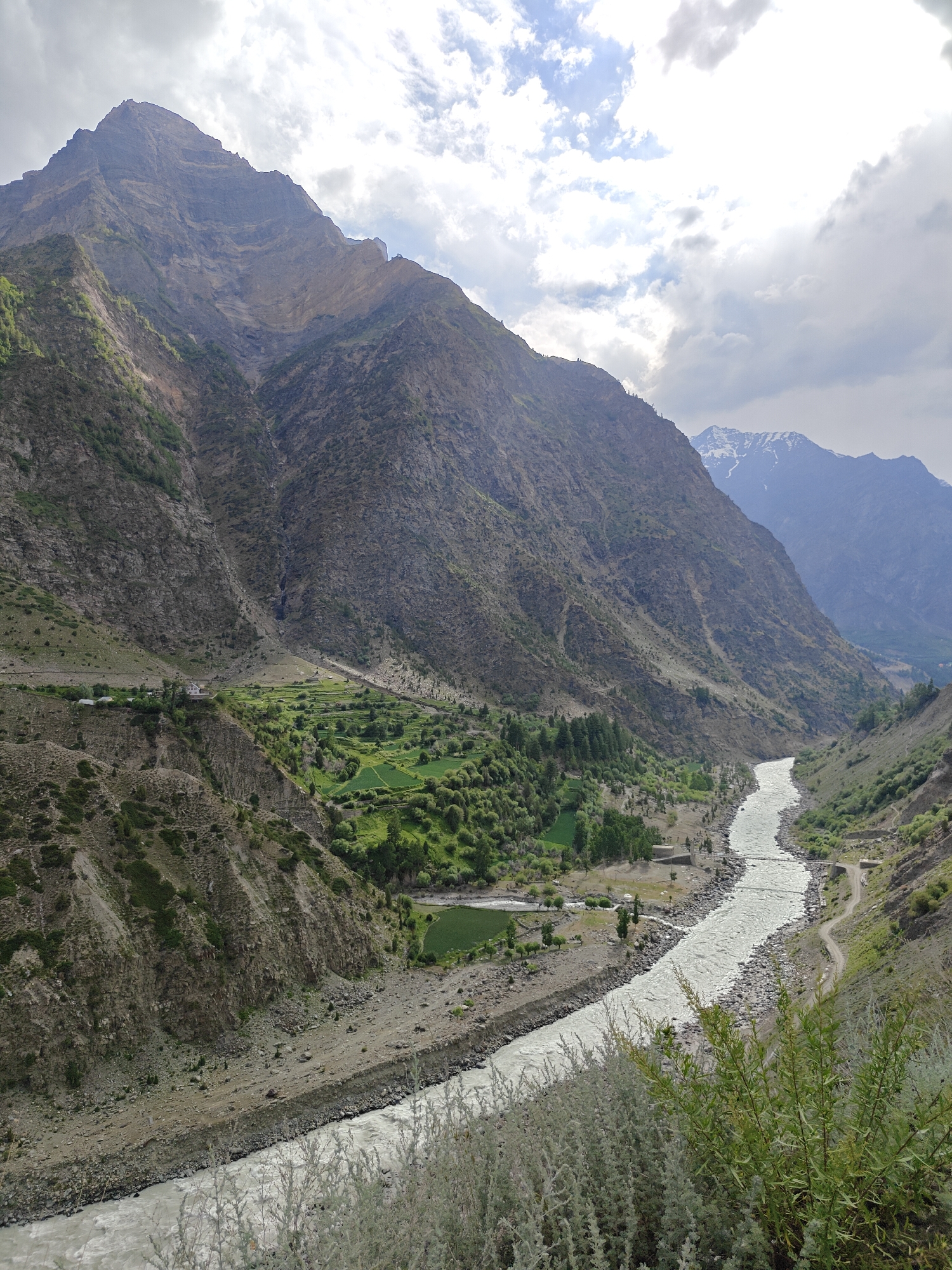 The image shows a wide, glacial river flowing through a valley in the Himalayas, likely near the village of Kirting, India. The river is a milky, white-grey color and appears to be fast-flowing, with visible rocks along its banks and bed. On either side of the river, steep, rocky mountains rise dramatically. The slopes are rugged, with patches of green vegetation and some sparse trees scattered across them.
In the mid-ground, on the left side of the valley, a small agricultural area is visible. These are terraced fields, cultivated with bright green crops, suggesting a fertile patch within the otherwise arid mountain landscape. A cluster of buildings, including a white house, is situated on a higher terrace, overlooking the fields and the river. A narrow road or track can be seen winding its way along the right bank of the river.
The sky above is partly cloudy, with dramatic formations of white and grey clouds against a bright sky, suggesting a dynamic weather system. The overall impression is one of natural grandeur, showcasing the raw beauty of a high-altitude river valley. The scene is tranquil and devoid of any human presence or activity beyond the cultivated fields and structures, emphasizing the vastness and power of nature.