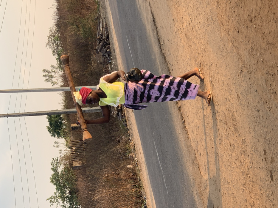 A woman in a yellow top, patterned skirt, and red hat carries a large wooden object on her shoulders while walking along a road. A small child is strapped to her back. She wears flip-flops. The road is paved on one side and dirt on the other. Vegetation, including trees and brush, borders the road. Utility poles are visible in the background.
