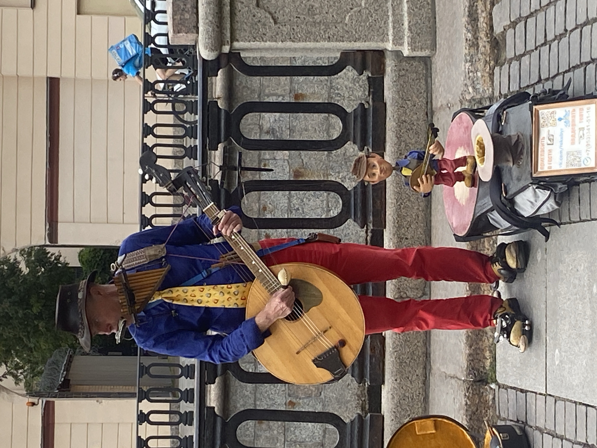 A street performer in Saint Petersburg, Russia, stands on a cobblestone sidewalk playing a lyre-guitar and a harmonica simultaneously. He wears a blue shirt, a yellow tie with a pattern, red trousers, a brown hat, and unique shoes. In front of him, a marionette in a brown hat and blue shirt holds a small guitar. A black railing separates the sidewalk from a canal or river. In the background, a person on a bicycle with a blue backpack rides past. The scene appears to be in a tourist area, given the presence of a performer and the architectural style.