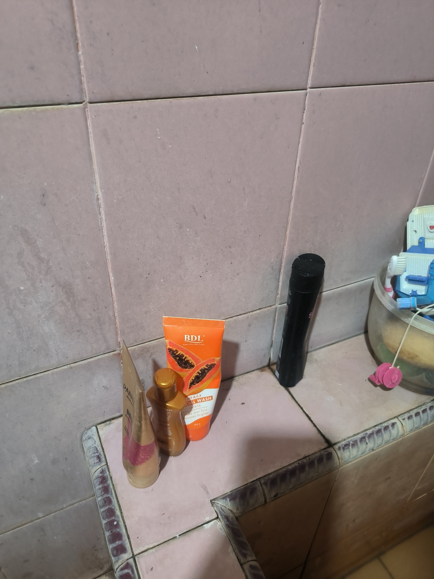 The image depicts a bathroom sink area with several personal care items. On the tiled surface, there are three bottles: a tall black bottle, a golden-brown bottle, and an orange tube labeled "BDL FACIAL WASH" with an image of papayas. To the left, a gold-colored tube is partially visible. In the background, a translucent plastic container holds toothbrushes and other toiletries, including a pink, spherical bath accessory. The tiles are a light pinkish-beige color, showing signs of wear and some discoloration. The overall environment appears to be a residential bathroom, likely within the city of Bumi Mulyo, Indonesia, given the prompt.