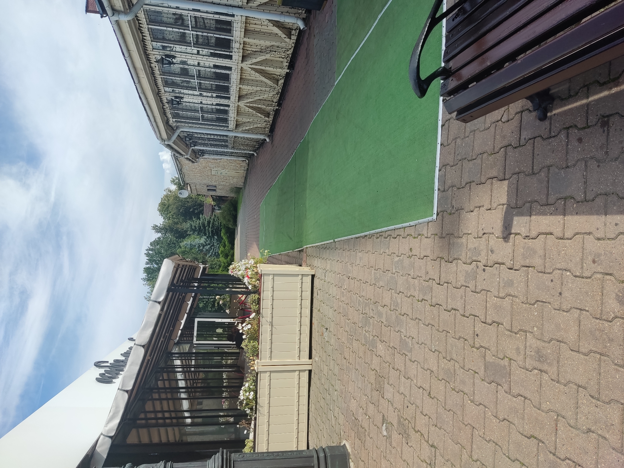 This is a ground-level, outdoor, vertical, full-shot image of a walkway next to a building in Noginsk, Russia. The walkway is paved with interlocking concrete pavers. To the left of the walkway is a building with a white facade and a covered outdoor seating area enclosed by glass. A long, beige planter box filled with white and pink flowers sits in front of the covered seating area. To the right of the walkway is a long, green carpet or artificial turf mat. The building to the right of the green mat has a stone facade and a light-colored, decorative wooden facade with windows. A black, wooden bench with ornate black metal armrests is positioned on the pavers to the right of the green mat, facing the building on the right. Trees and a blue sky with white clouds are visible in the background. No people are visible in the image.