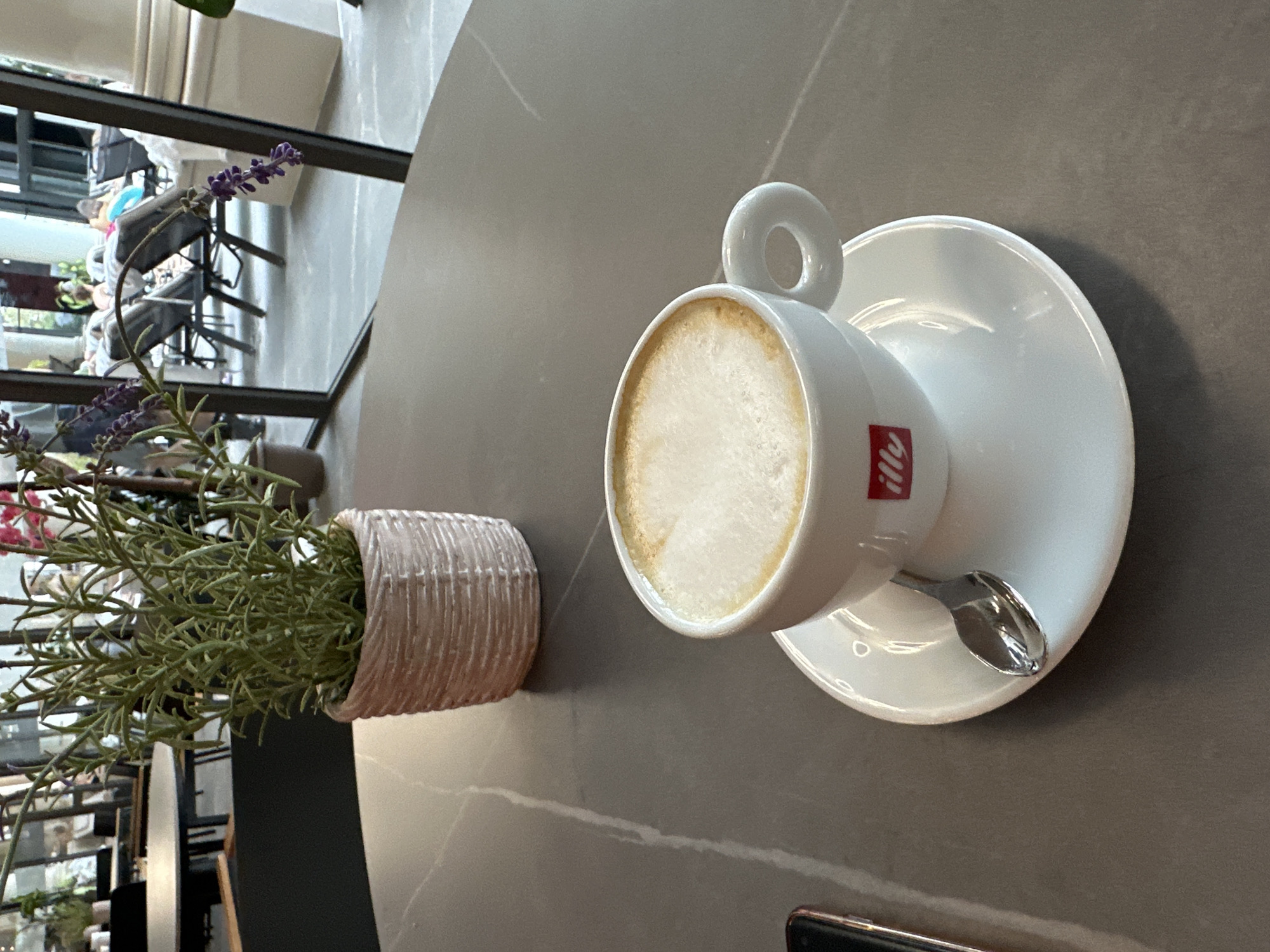 The image depicts a coffee cup and saucer on a table, next to a potted plant with purple flowers. The cup is white with a red logo. There is a silver spoon resting on the saucer. The table is gray and appears to be in a cafe or restaurant setting. Based on the "Illy" logo on the cup, the photo was likely taken at Illy, which is located in Kazanlak, Bulgaria.