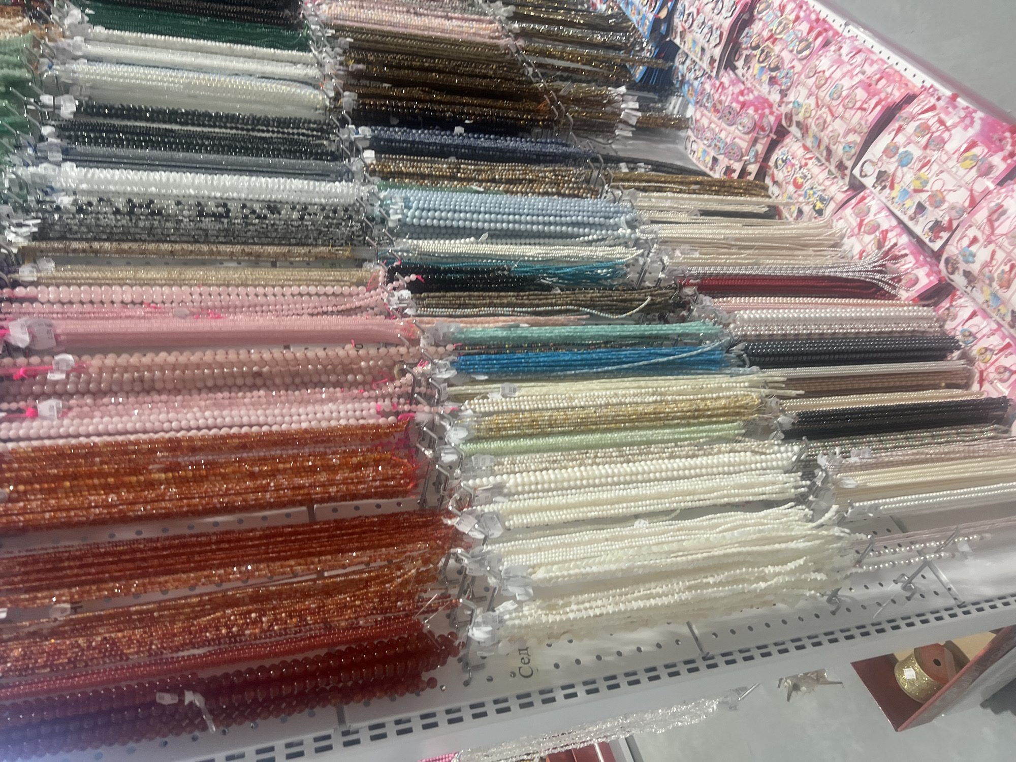 The image shows a retail display of various beaded strands arranged on shelves. The beads come in a range of colors including green, white, black, gray, gold, pink, orange, red, blue, and beige. The display is housed in a store located in Burgas, Bulgaria. Other accessories are located along the right side.