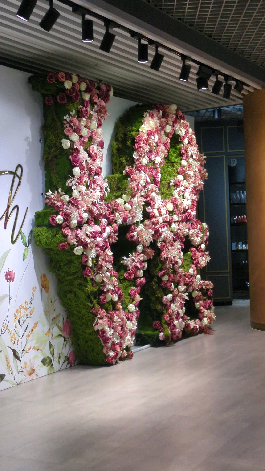 The image shows a decorative display of two large letters, possibly an "H", made of green moss and covered with numerous pink and white roses. The letters are positioned against a white wall with a floral illustration. Above the display, a series of black track lights are visible, illuminating the scene. In the background, a dark blue cabinet with glass doors and shelves can be seen, containing various items. A light brown cylindrical pillar is to the right of the display. The floor is a light-colored, smooth surface. The overall impression is one of a festive or elegant event setting. The image does not provide any specific information about the city of Chișinău, Moldova.