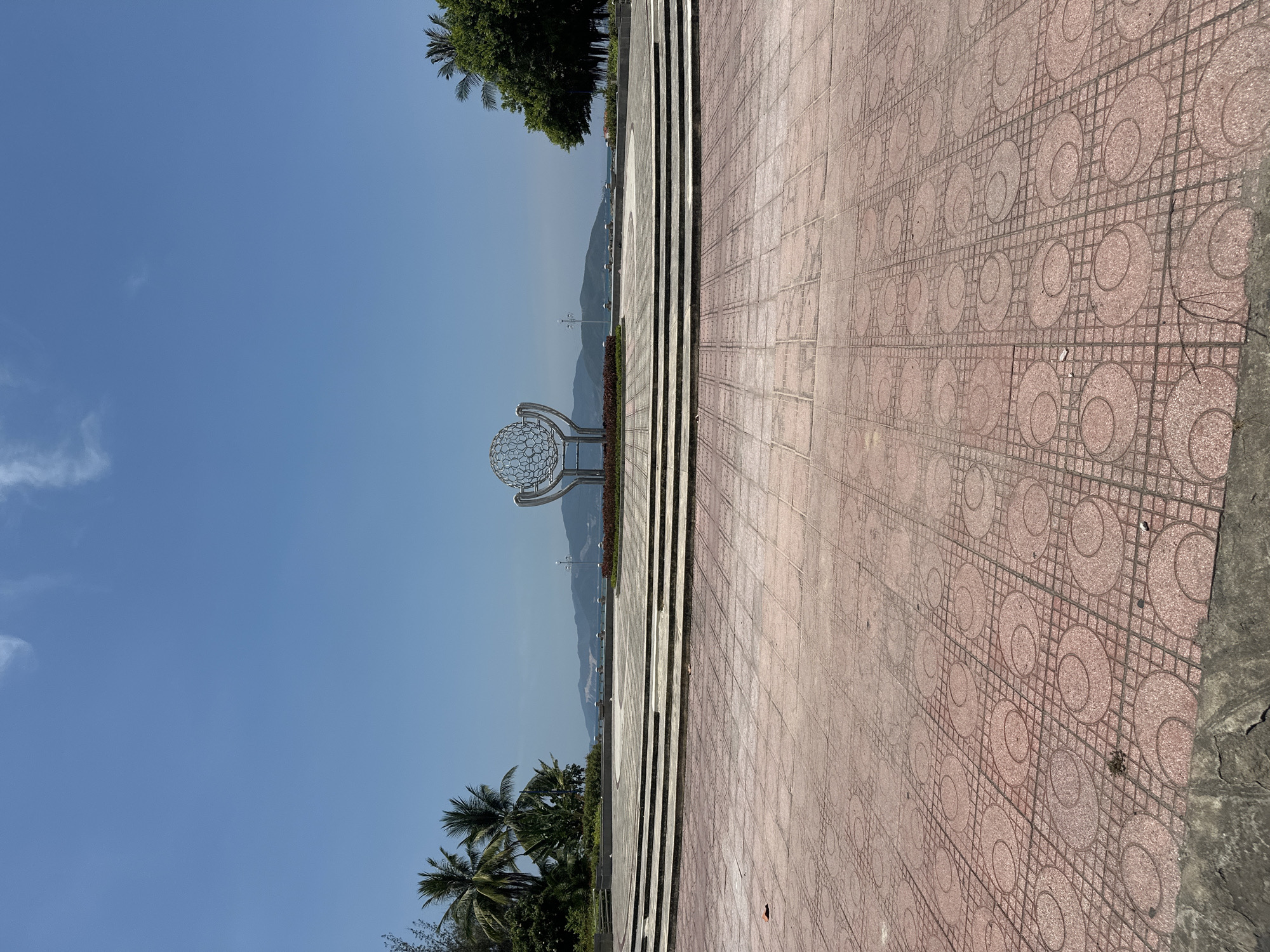 The image shows a wide tiled public space in Bac Nha Trang Ward, Vietnam. The tiles are pink with a circular pattern. In the background, there is a metal globe-shaped sculpture, mountains, and a clear blue sky. There are trees along the left edge of the image. Steps lead towards the sculpture. No people are visible. The photo is taken during daylight.