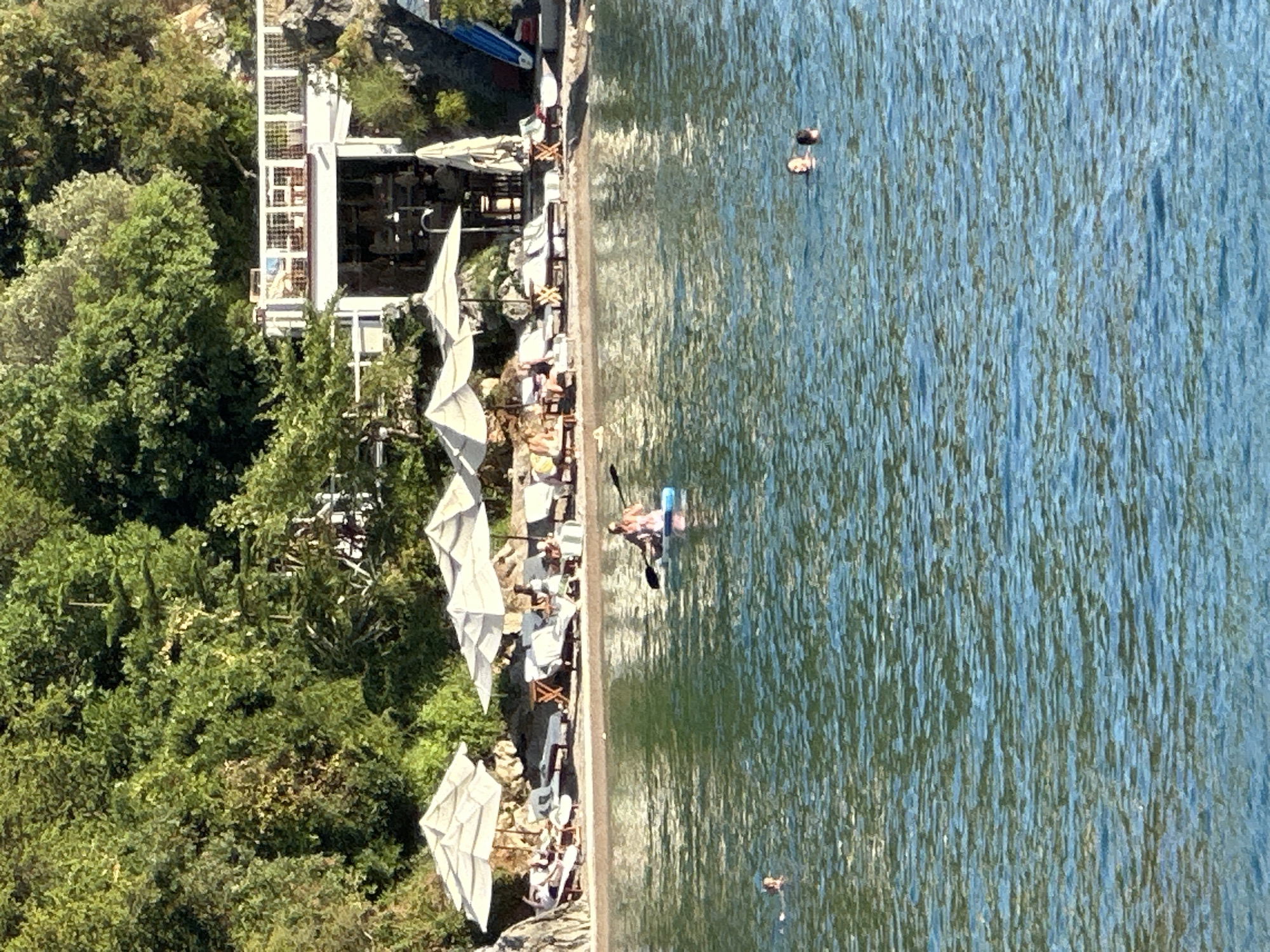 The image depicts a sunny day at a beach resort area, likely near Budva, Montenegro, characterized by lush green trees and rocky terrain rising behind a beach. The beach itself features numerous lounge chairs and umbrellas, with several people relaxing or sunbathing. The water in the foreground is clear and blue, with ripples on its surface. In the water, one person is paddleboarding, while two others are swimming. A building, possibly a restaurant or hotel, is visible in the background, perched on a higher elevation and overlooking the beach and water. The scene suggests a relaxed vacation atmosphere.